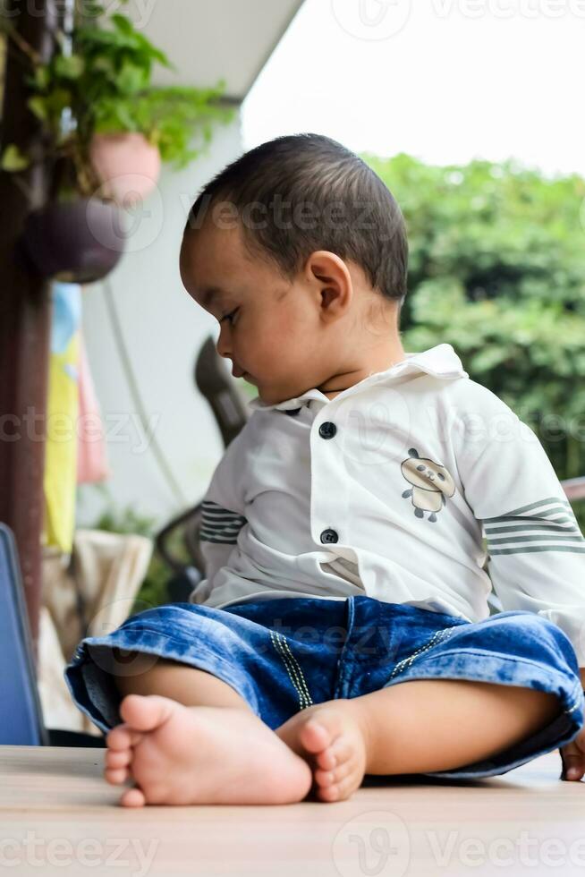 bebé chico a hogar balcón. brillante retrato de contento niño sentado en el mesa. pequeño 1 año antiguo chico durante el día ligero a casa balcón. foto