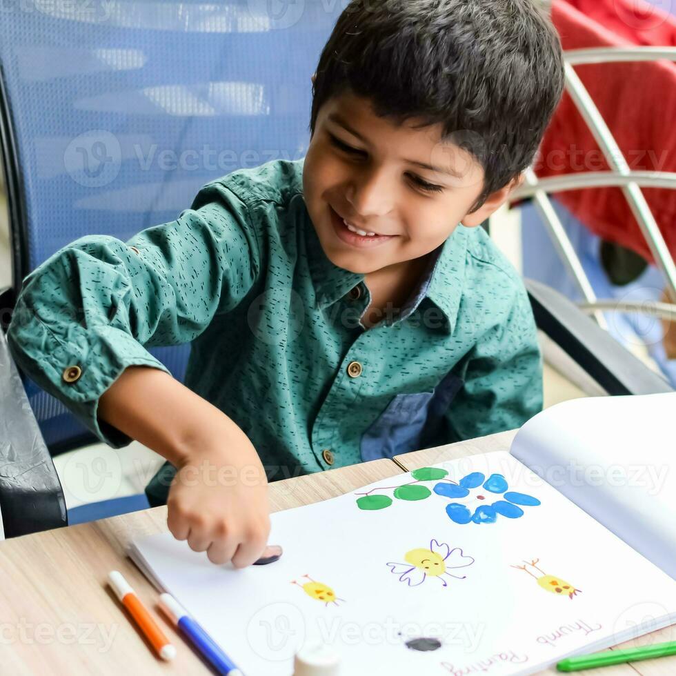 Smart Indian little boy perform thumb painting with different colourful water colour kit during the summer vacations, Cute Indian Kid doing colourful thumb painting drawing on wooden table photo