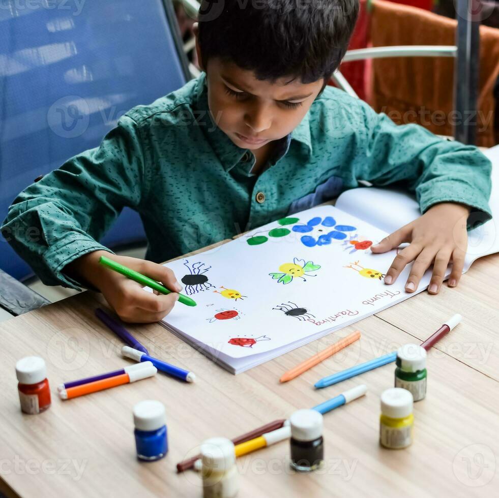 Smart Indian little boy perform thumb painting with different colourful water colour kit during the summer vacations, Cute Indian Kid doing colourful thumb painting drawing on wooden table photo