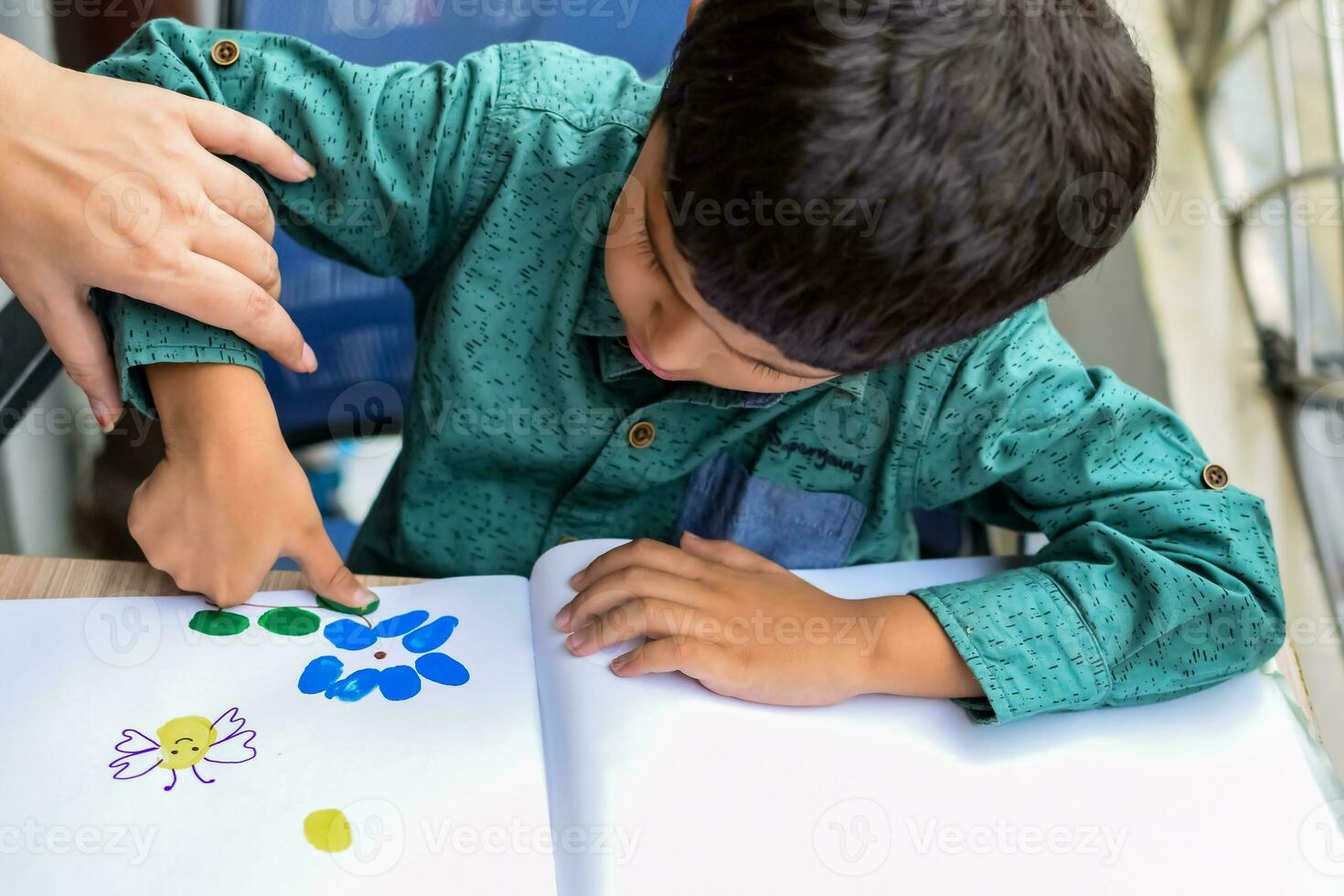 Smart Indian little boy perform thumb painting with different colourful water colour kit during the summer vacations, Cute Indian Kid doing colourful thumb painting drawing on wooden table photo