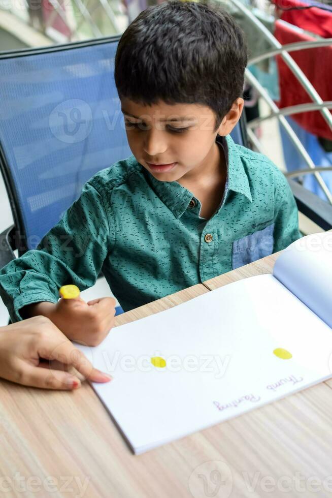 Smart Indian little boy perform thumb painting with different colourful water colour kit during the summer vacations, Cute Indian Kid doing colourful thumb painting drawing on wooden table photo