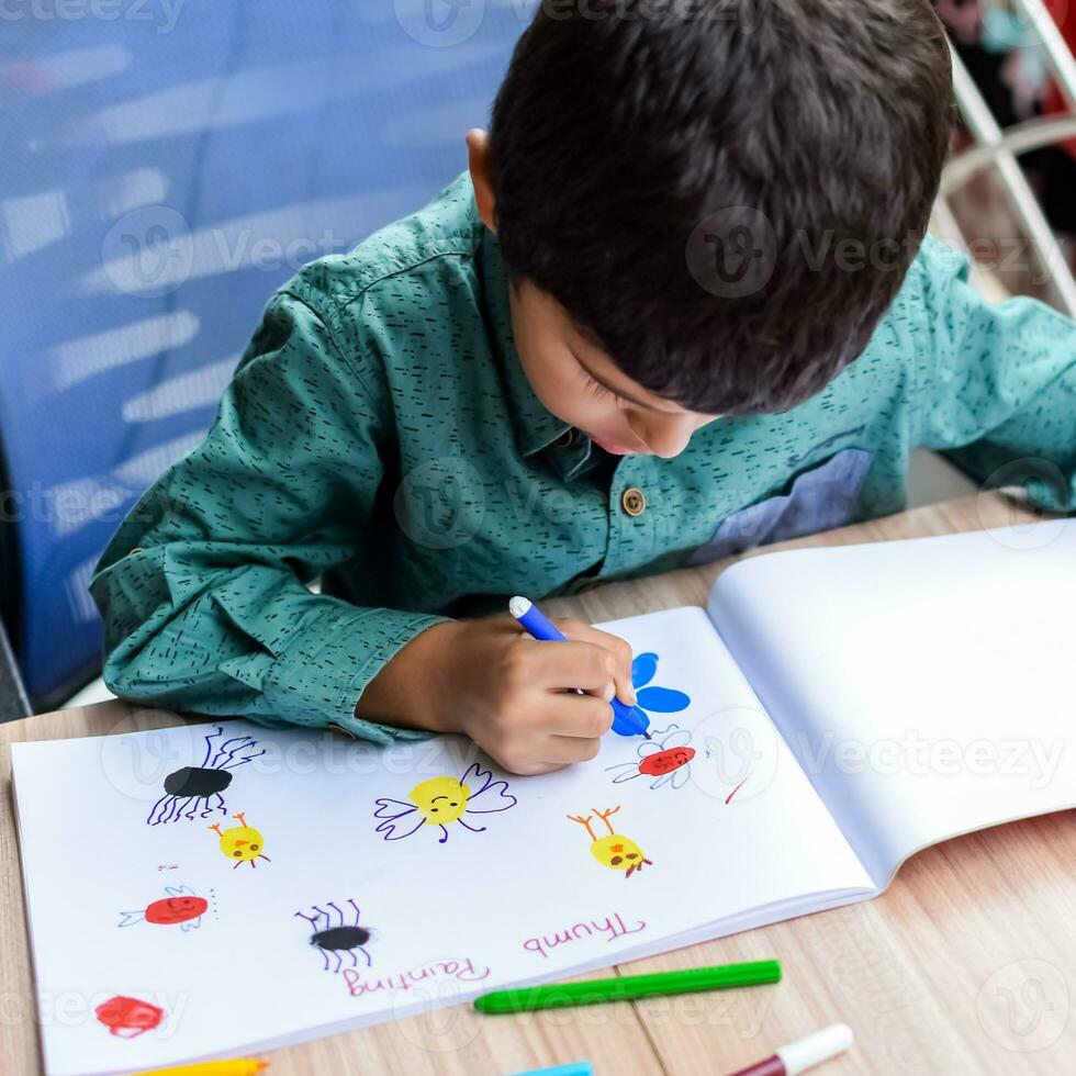 Smart Indian little boy perform thumb painting with different colourful water colour kit during the summer vacations, Cute Indian Kid doing colourful thumb painting drawing on wooden table photo