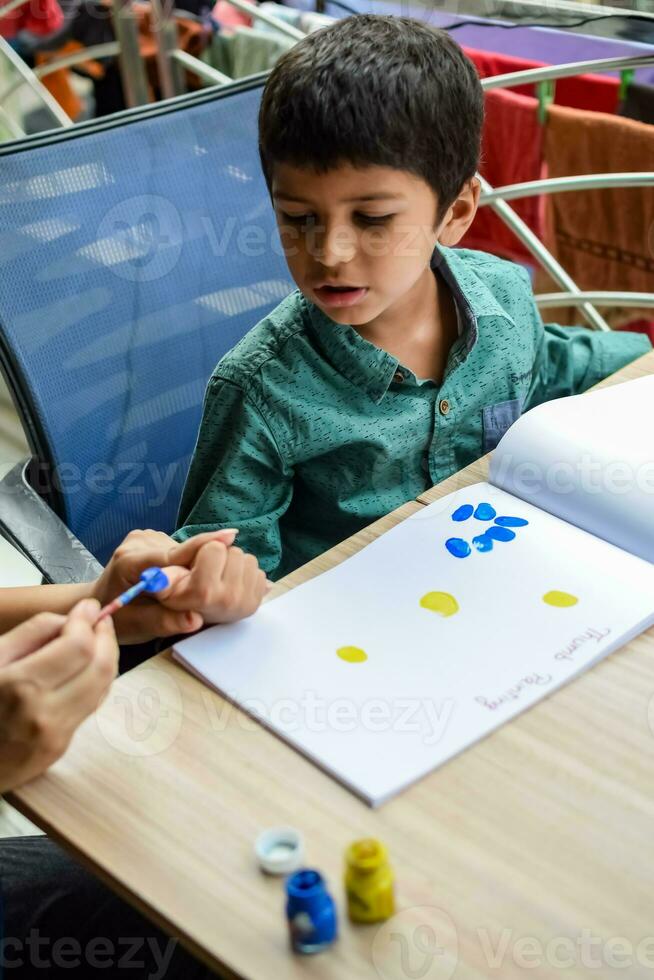 Smart Indian little boy perform thumb painting with different colourful water colour kit during the summer vacations, Cute Indian Kid doing colourful thumb painting drawing on wooden table photo