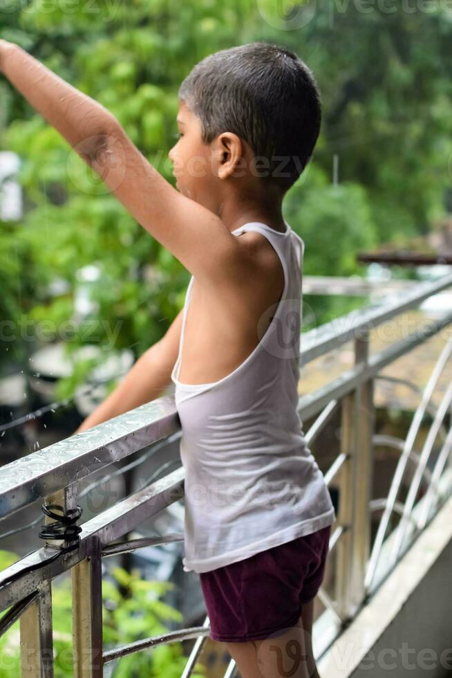 pequeño niño jugando en verano lluvia en casa balcón, indio inteligente chico jugando con lluvia gotas durante monzón lluvioso estación, niño jugando en lluvia foto