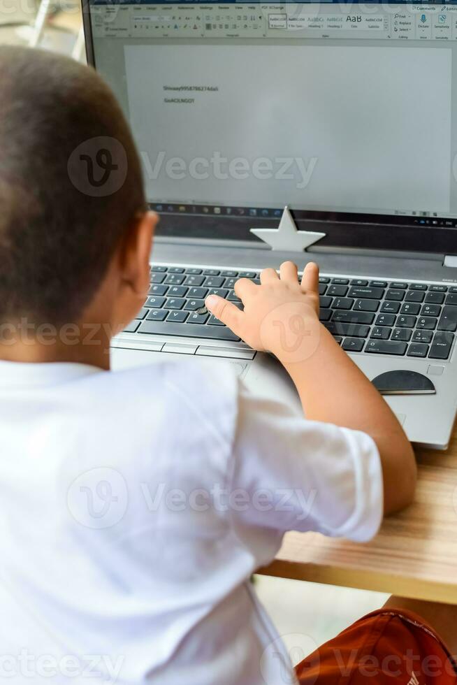 pequeño chico sentado a mesa utilizando ordenador portátil para en línea clase en grado 1, niño estudiando en ordenador portátil desde hogar para distancia aprendizaje en línea educación, colegio chico niños estilo de vida concepto foto