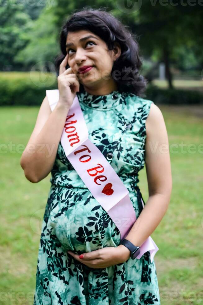 un embarazada indio dama poses para al aire libre el embarazo disparar y manos en barriga, indio embarazada mujer pone su mano en su estómago con un maternidad vestir a sociedad parque, embarazada fuera de maternidad disparar foto