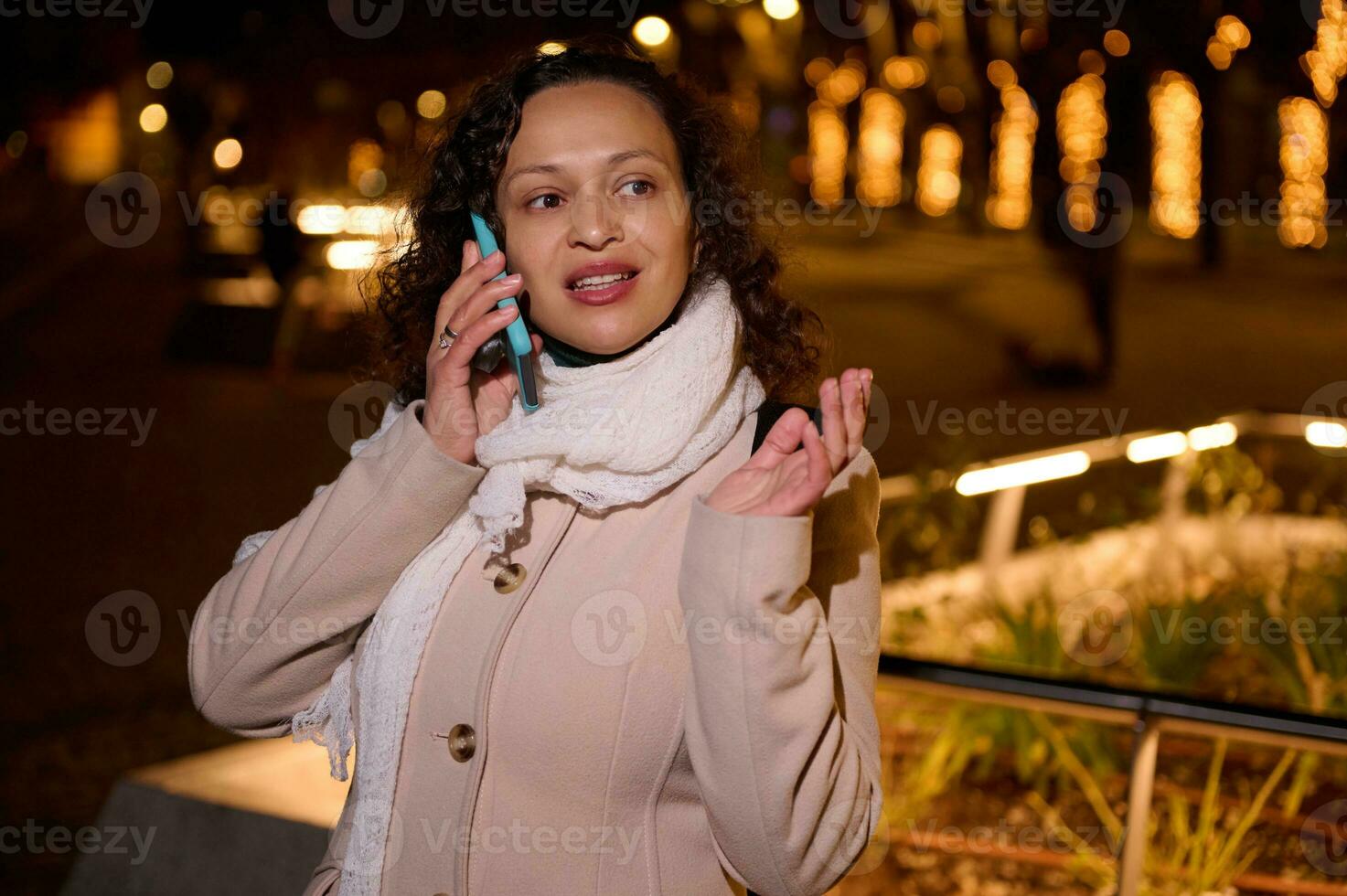 multi étnico joven adulto mujer sonrisas mientras negociaciones en móvil teléfono, en pie en el iluminado ciudad calle por noche foto