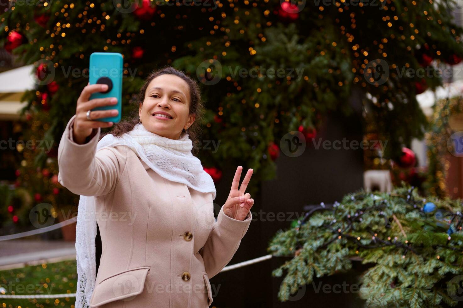 Beautiful woman stands against Xmas tree decorated with garlands, shows peace sign while makes selfie at Christmas fair photo