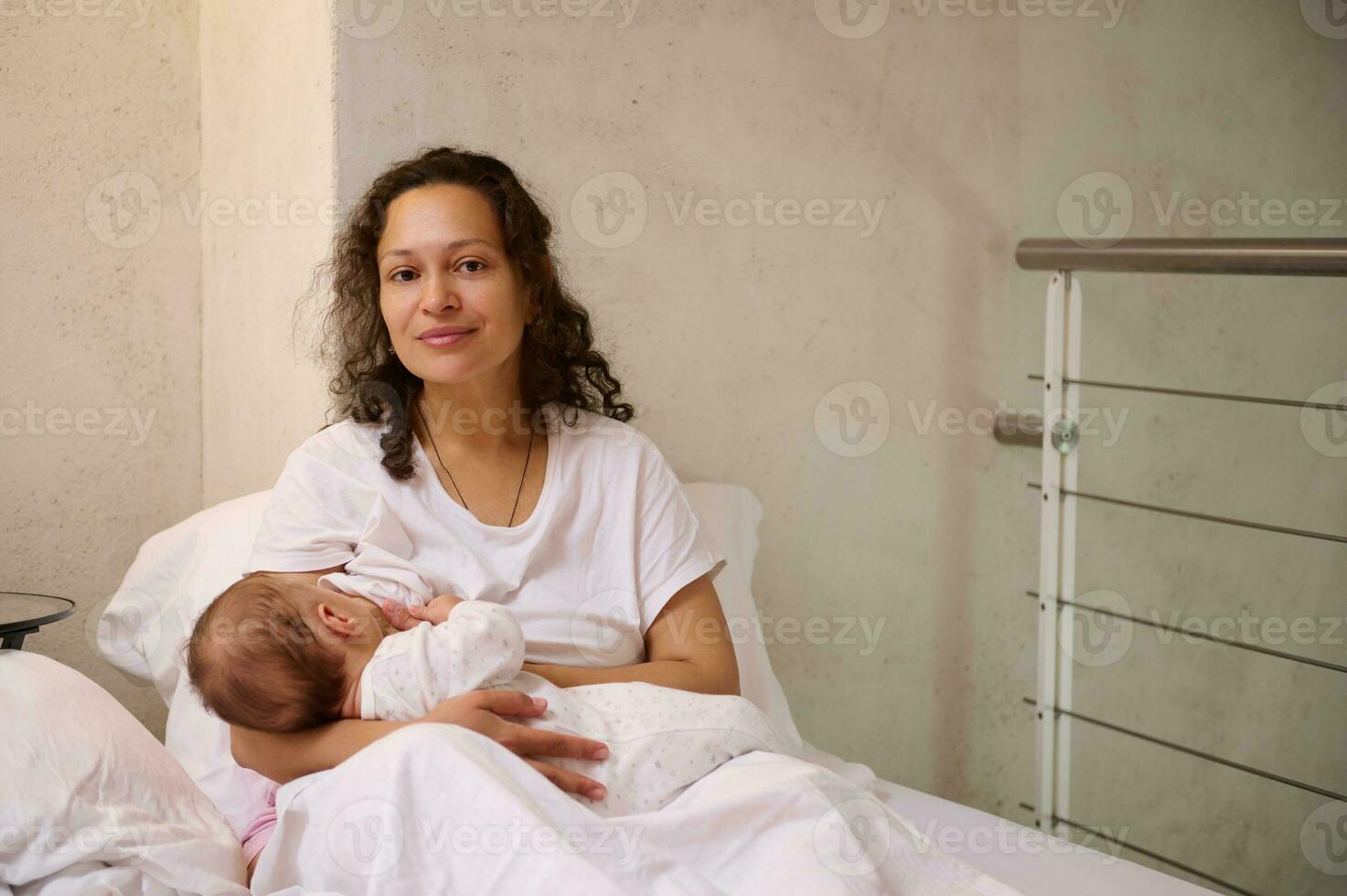 Beautiful Latin American woman, young loving caring mother in white pajamas, breastfeeding her baby in cozy home bedchamber, smiling and confidently looking at the camera. Maternity leave lifestyle photo