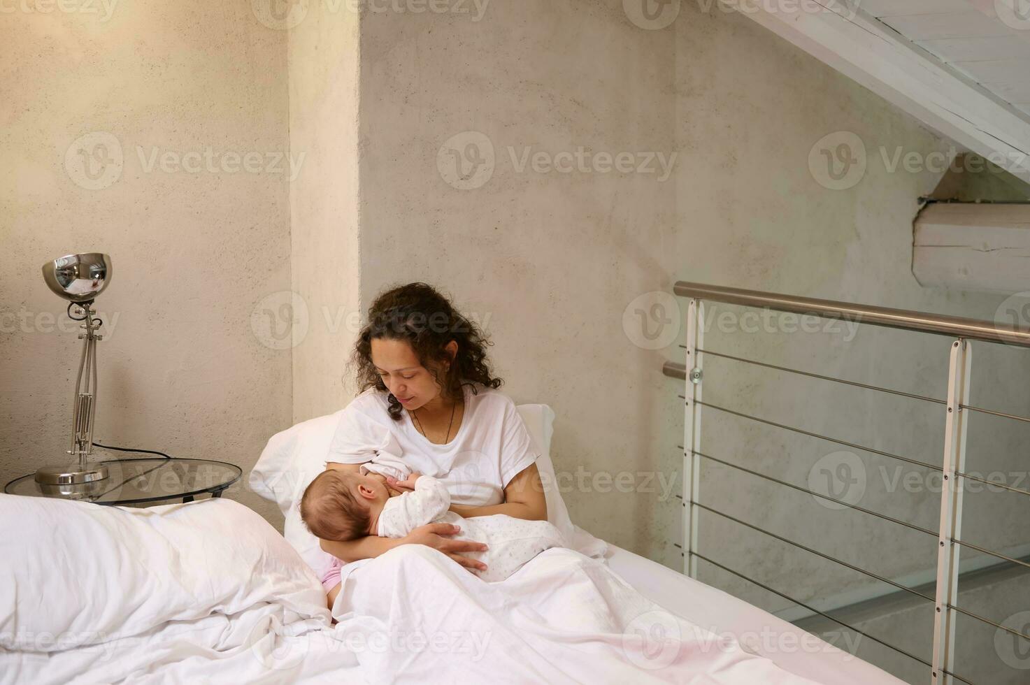 Young woman mother holding and breastfeeding her newborn baby, sitting on the bed in white bedchamber interior photo