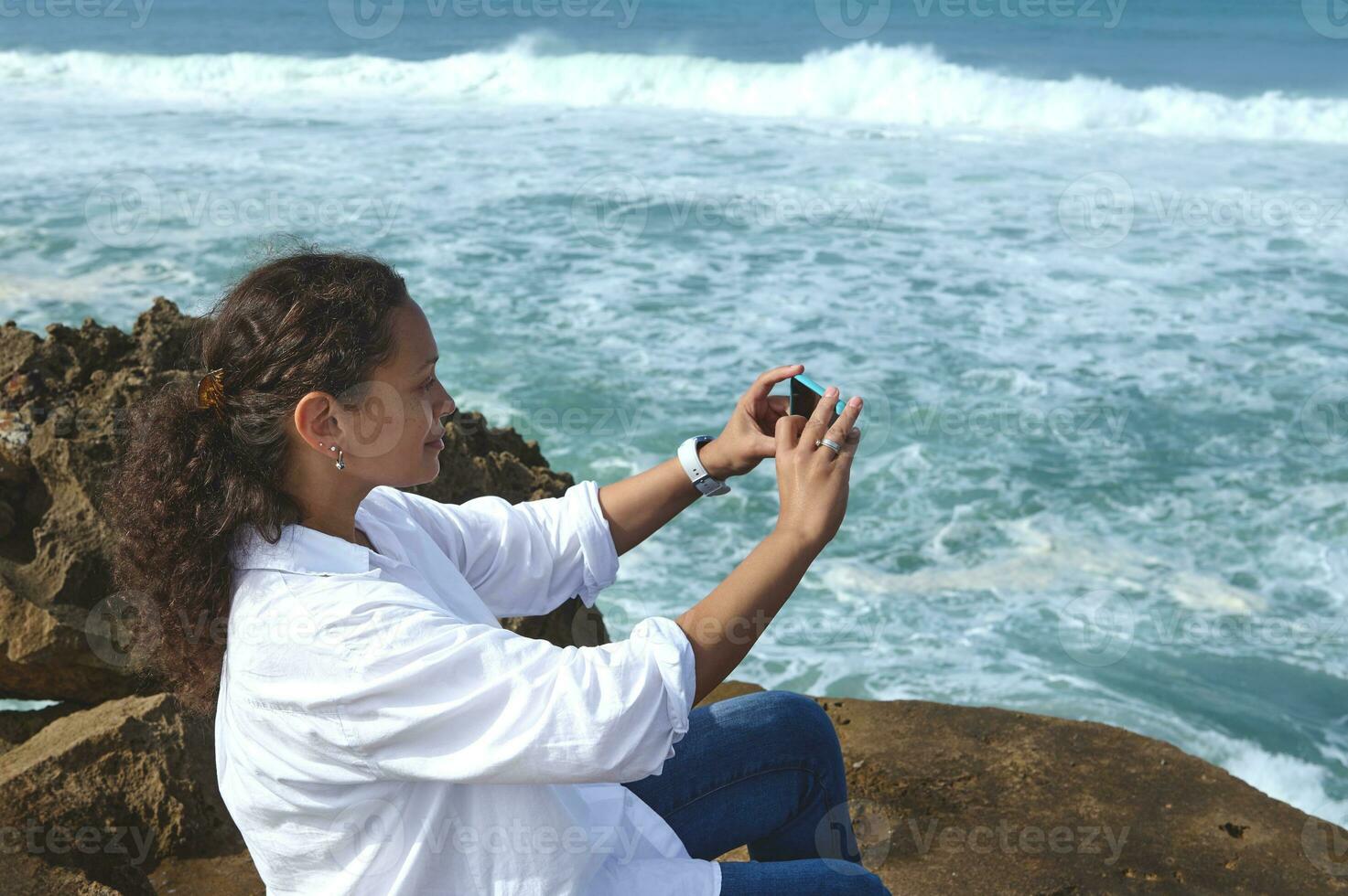 Curly multi ethnic pretty woman taking photo on her smart mobile phone, sitting on the rock on the Atlantic ocean beach