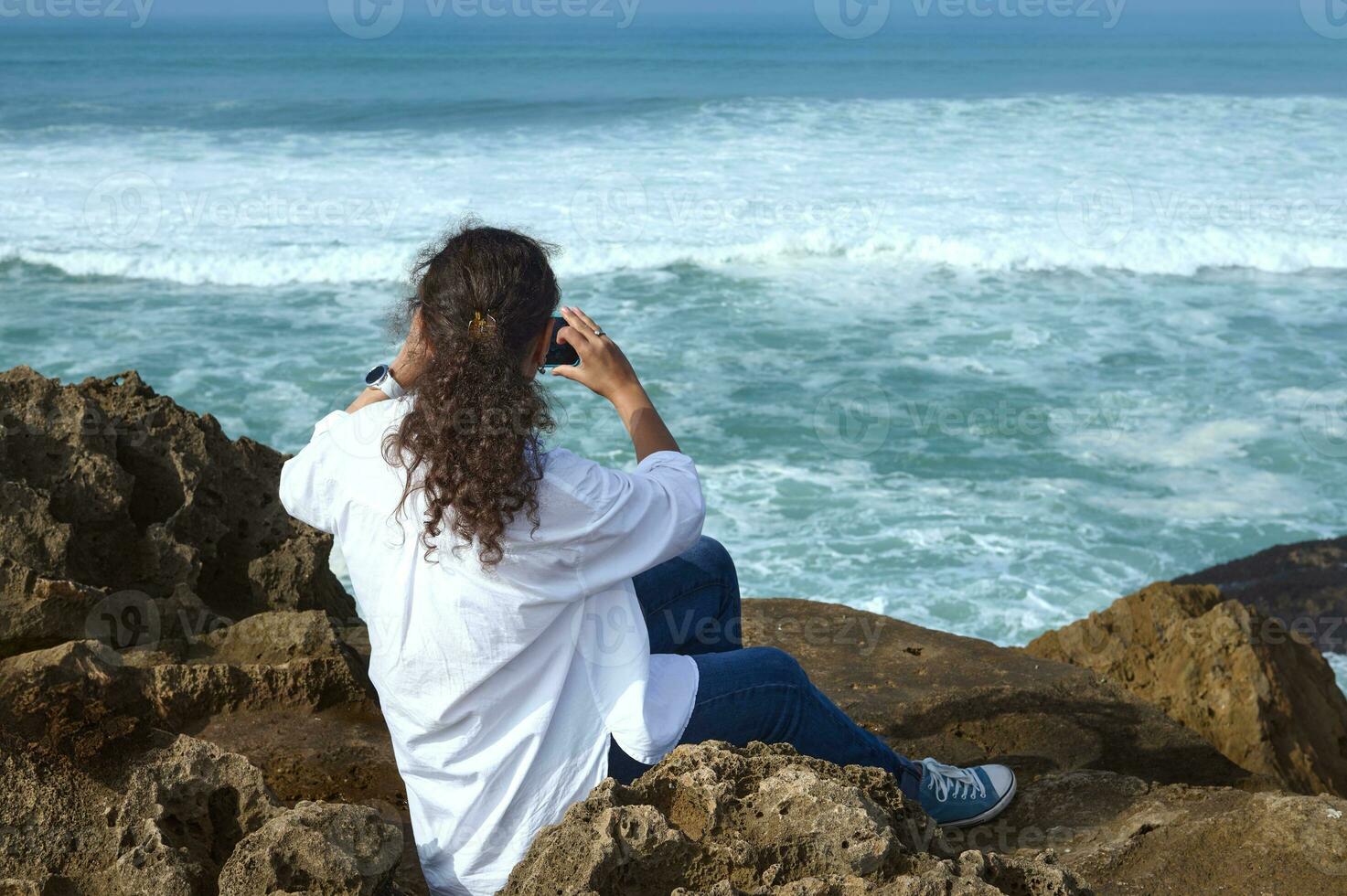 multi étnico bonito mujer tomando foto en su inteligente móvil teléfono, sentado en el rock en el atlántico Oceano playa