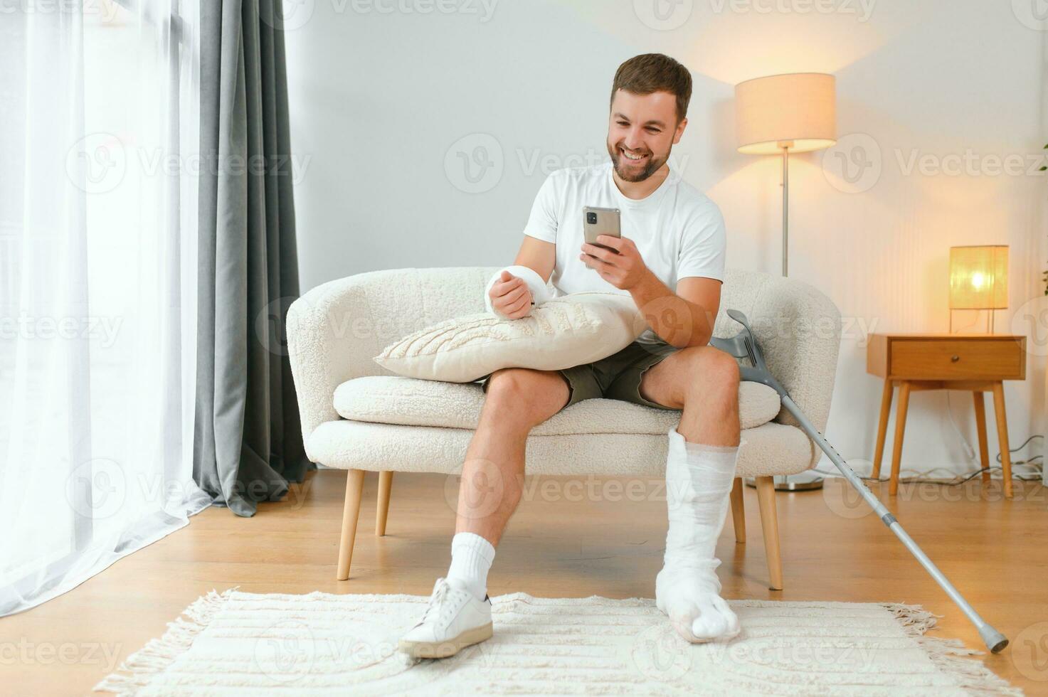 Man with a broken leg and arm using his mobile phone while relaxing on the sofa at home. Accident, injury, treatment, rehabilitation concept photo