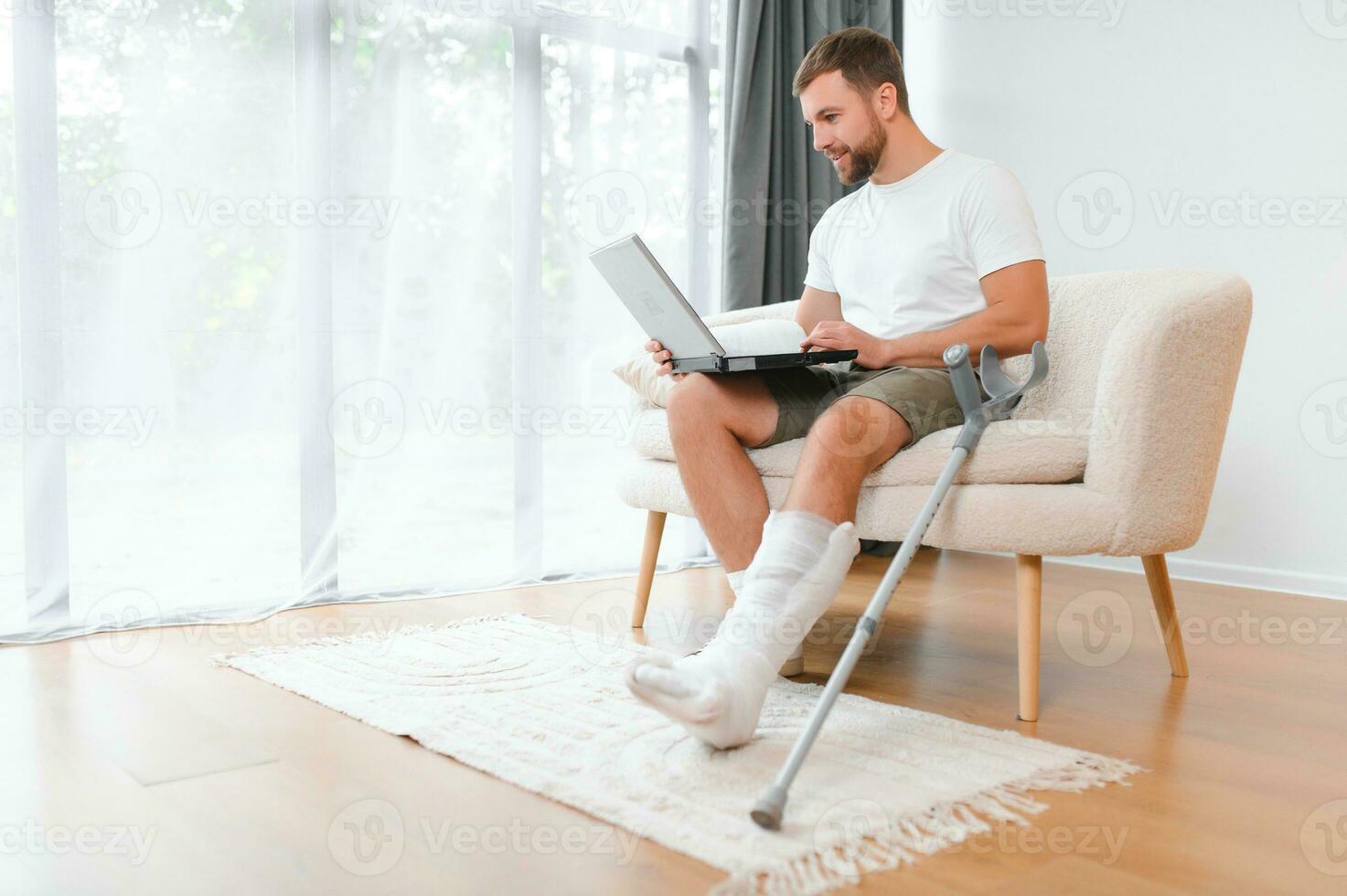 Happy young man with arm in a cast sitting on the couch at home and communicating on a laptop photo