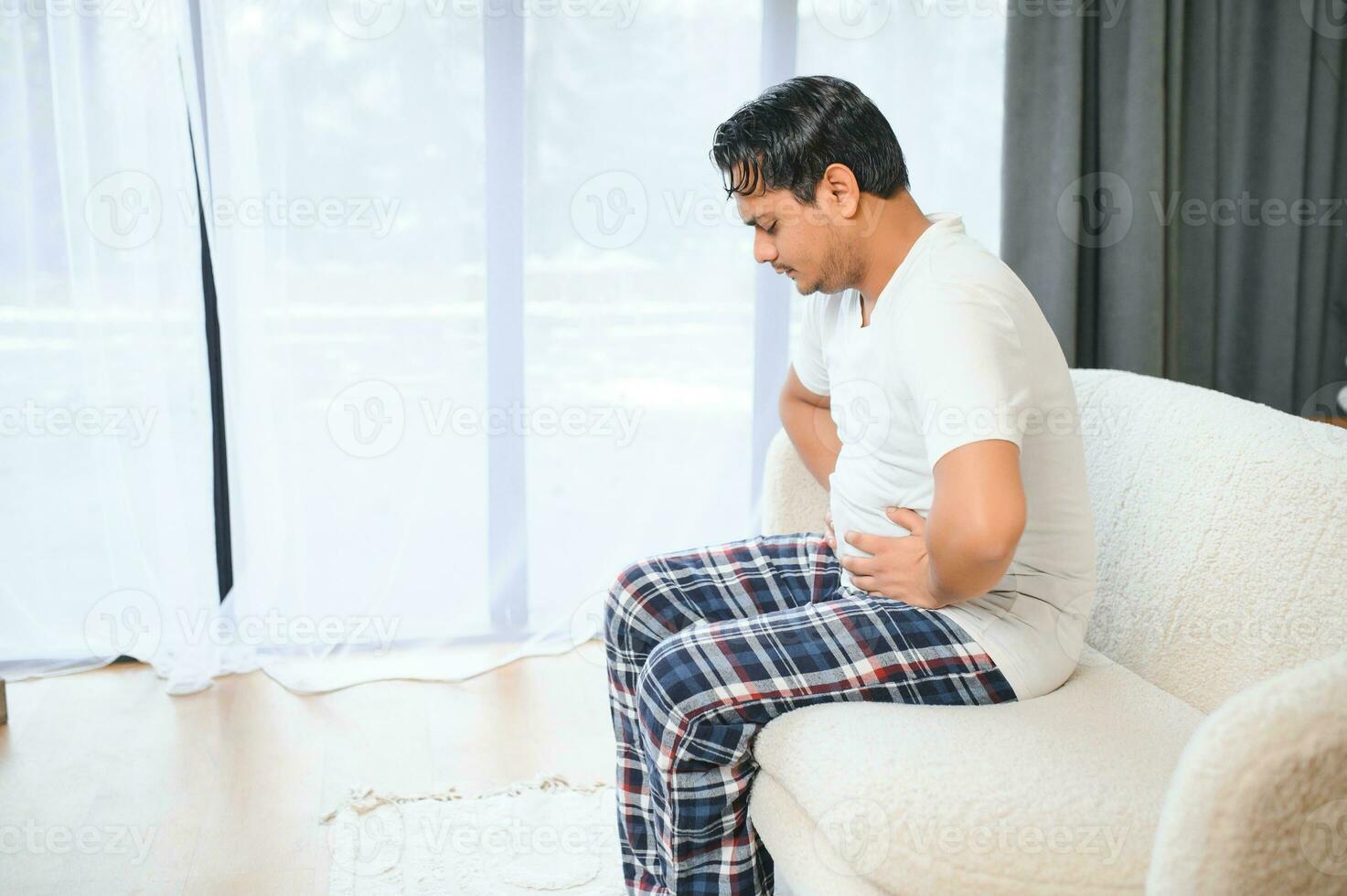 Unhappy indian or arabian man, sits on comfortable sofa in cozy living room, holds his hands on his stomach, grimaces from pain in his stomach, suffers from poisoning, spasm, stomach problems. photo