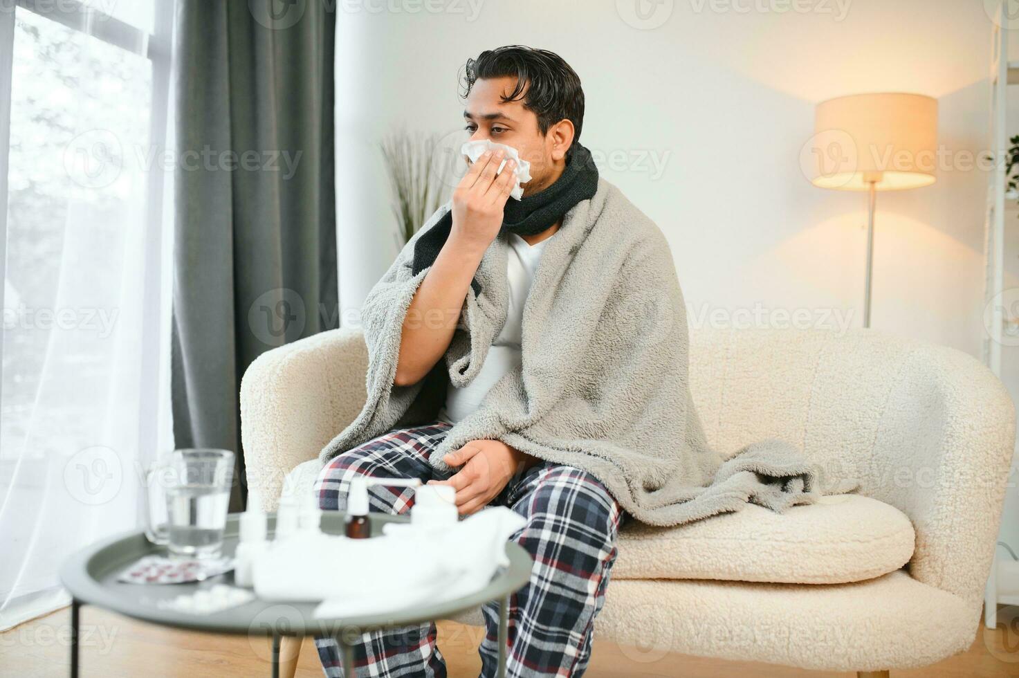 Sick man sitting at home on sofa in living room, sneezing has allergy and runny nose, Indian man with napkin has a cold photo