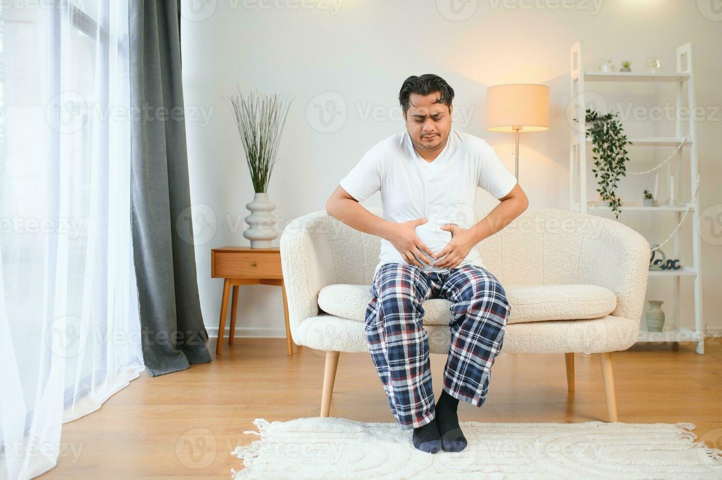 Unhappy indian or arabian man, sits on comfortable sofa in cozy living room, holds his hands on his stomach, grimaces from pain in his stomach, suffers from poisoning, spasm, stomach problems. photo