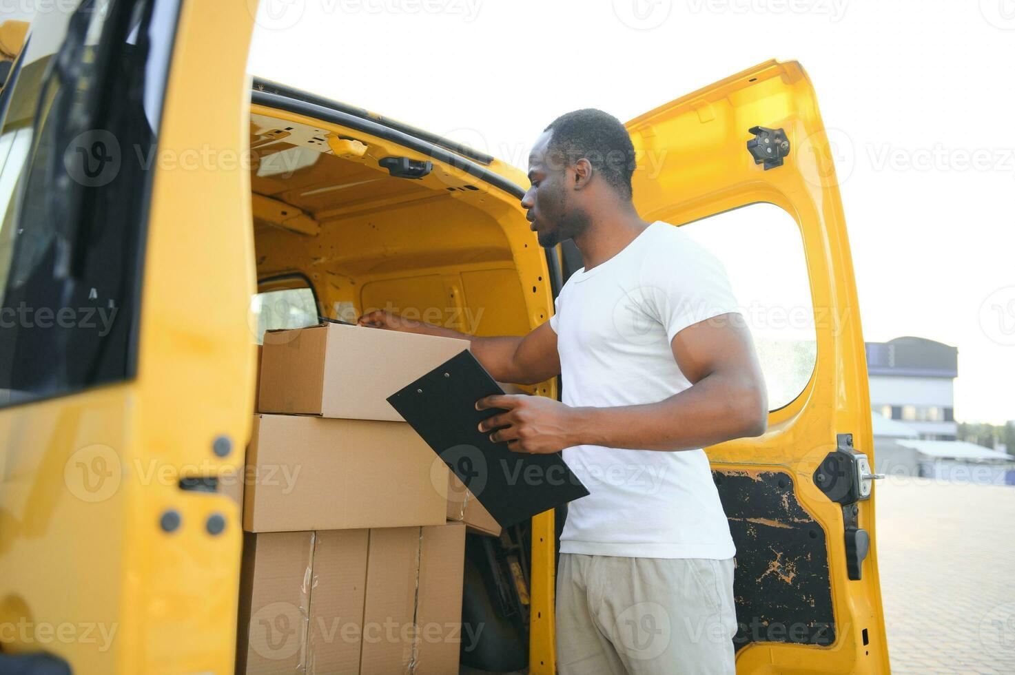 african american Worker unloading boxes from van outdoors. Moving service concept. Male employee of moving company makes inventory of packed boxes and writes data to clipboard. photo