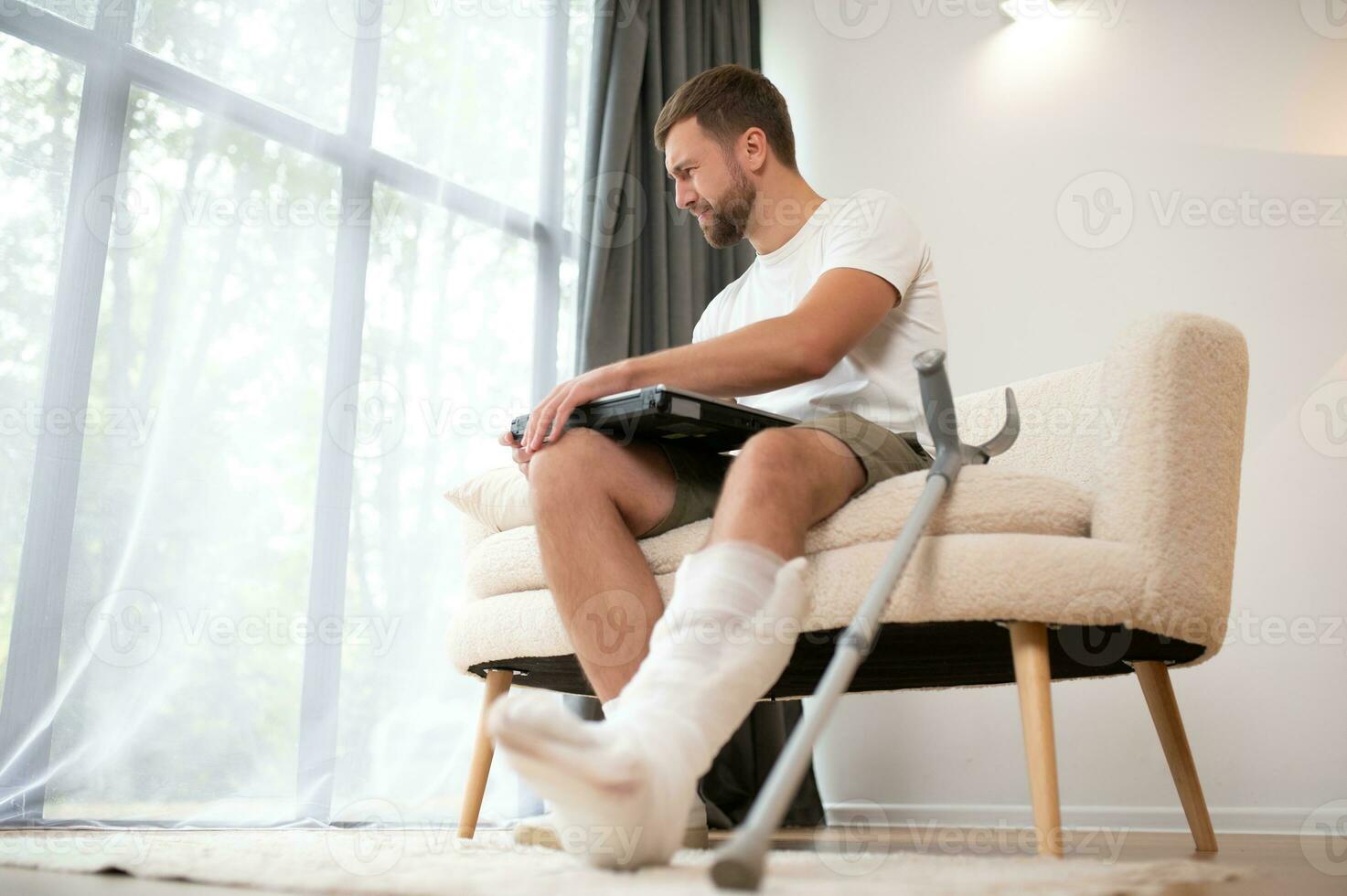 Happy young man with arm in a cast sitting on the couch at home and communicating on a laptop photo