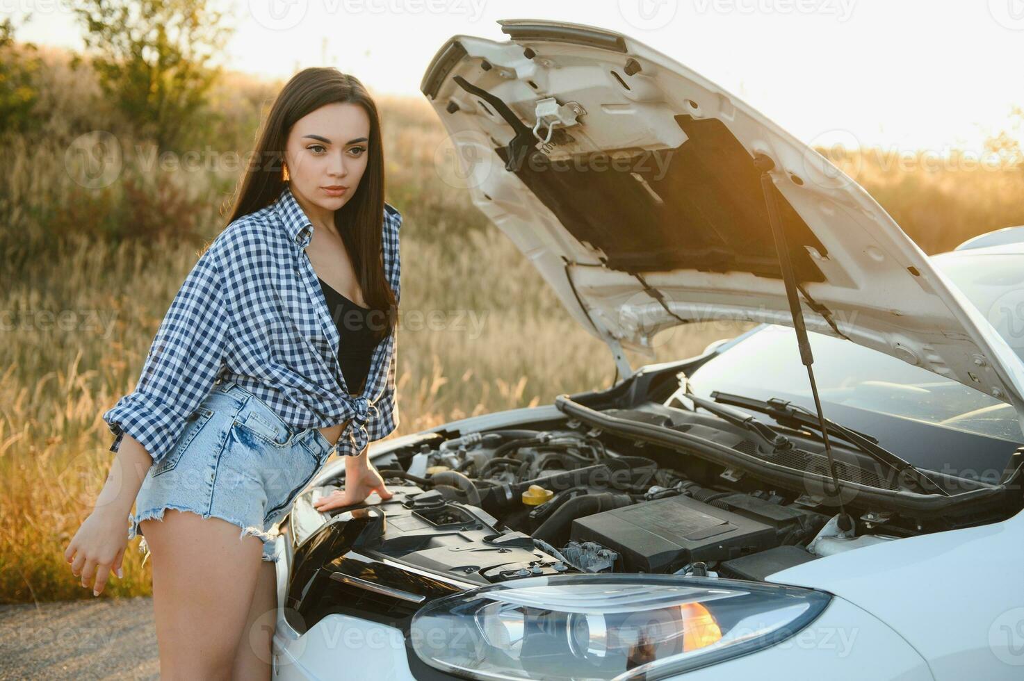 Attractive slim young girl in summer shorts and shirt repairs a broken car. A beautiful woman stands near raised car hood. photo