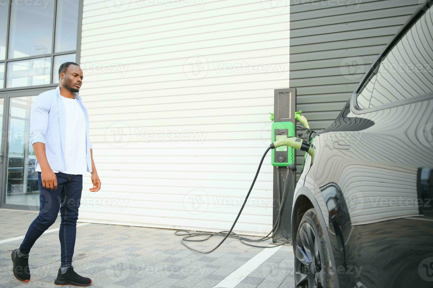 African man standing near electric car with charging cable in plug. Eco friendly vehicle charging on station. LIfestyles concept photo