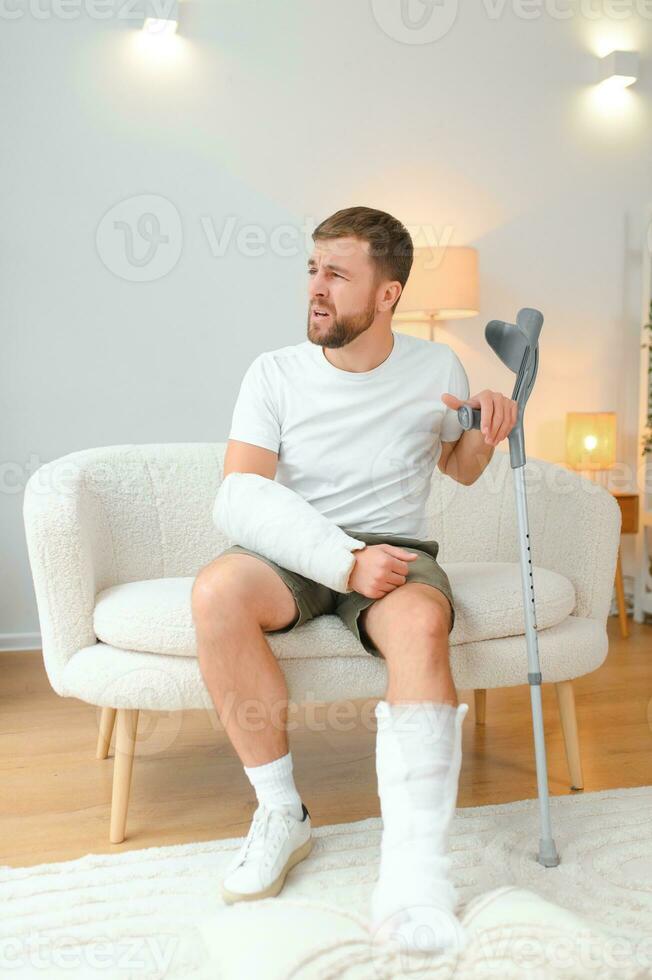 man recovery from accident fracture broken bone injury with leg splints in cast neck splints collar arm splints sling support arm in living room. Social security and health insurance concept photo