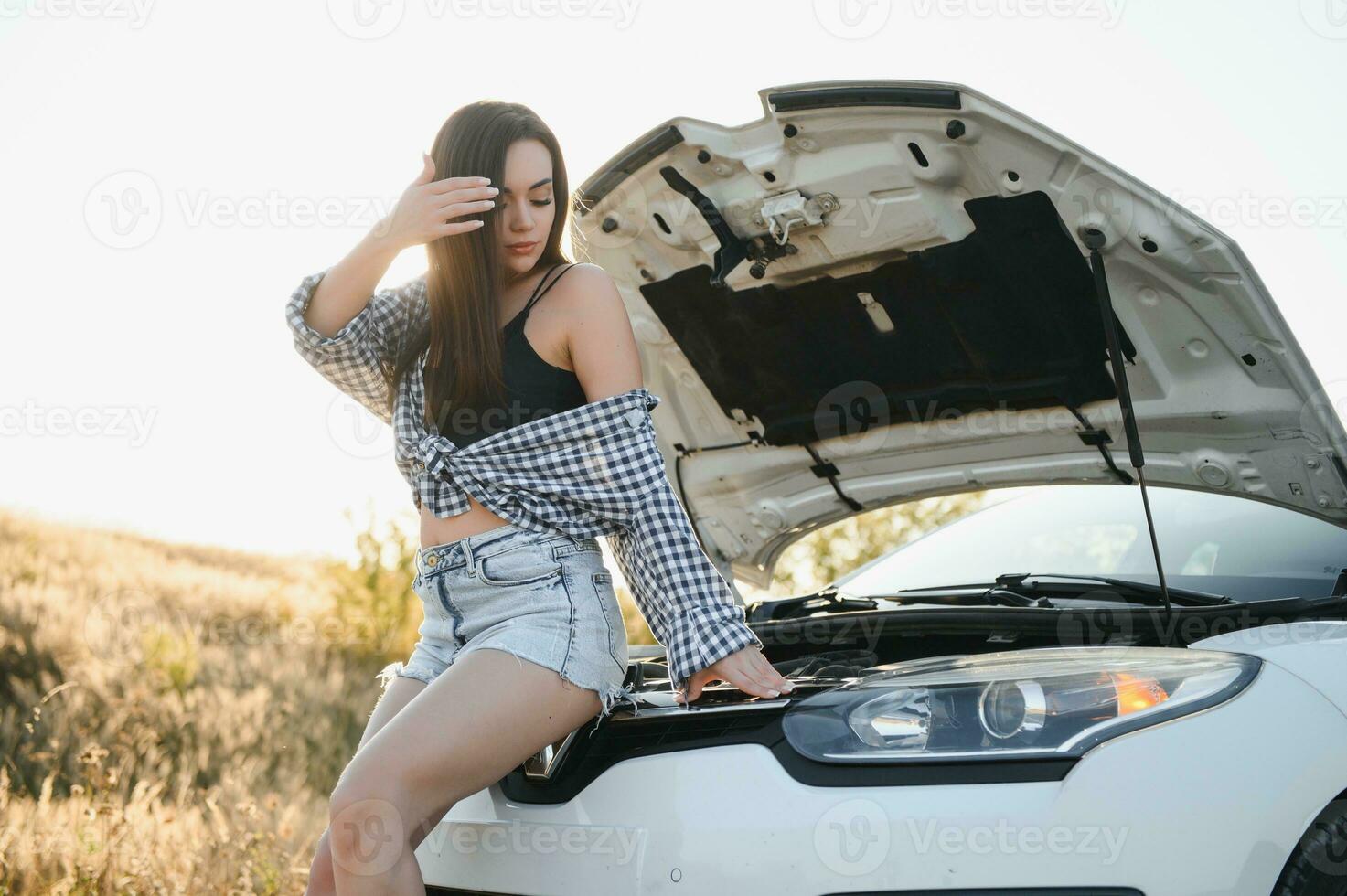 Beautiful slim girl in shirt and shorts looks in open car hood on a road photo