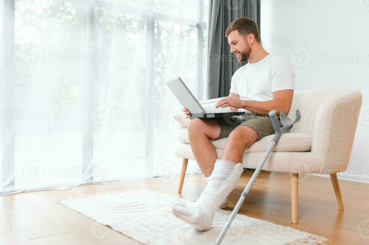 Happy young man with arm in a cast sitting on the couch at home and communicating on a laptop photo