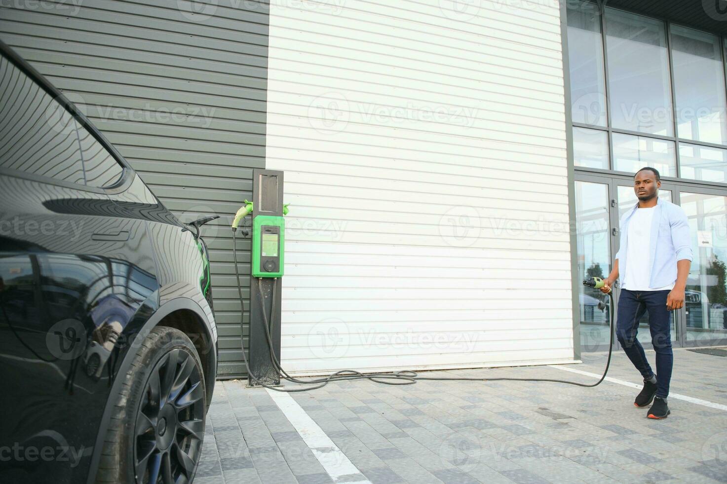 Serious african man holding charge cable in on hand, standing near luxury electric car. photo