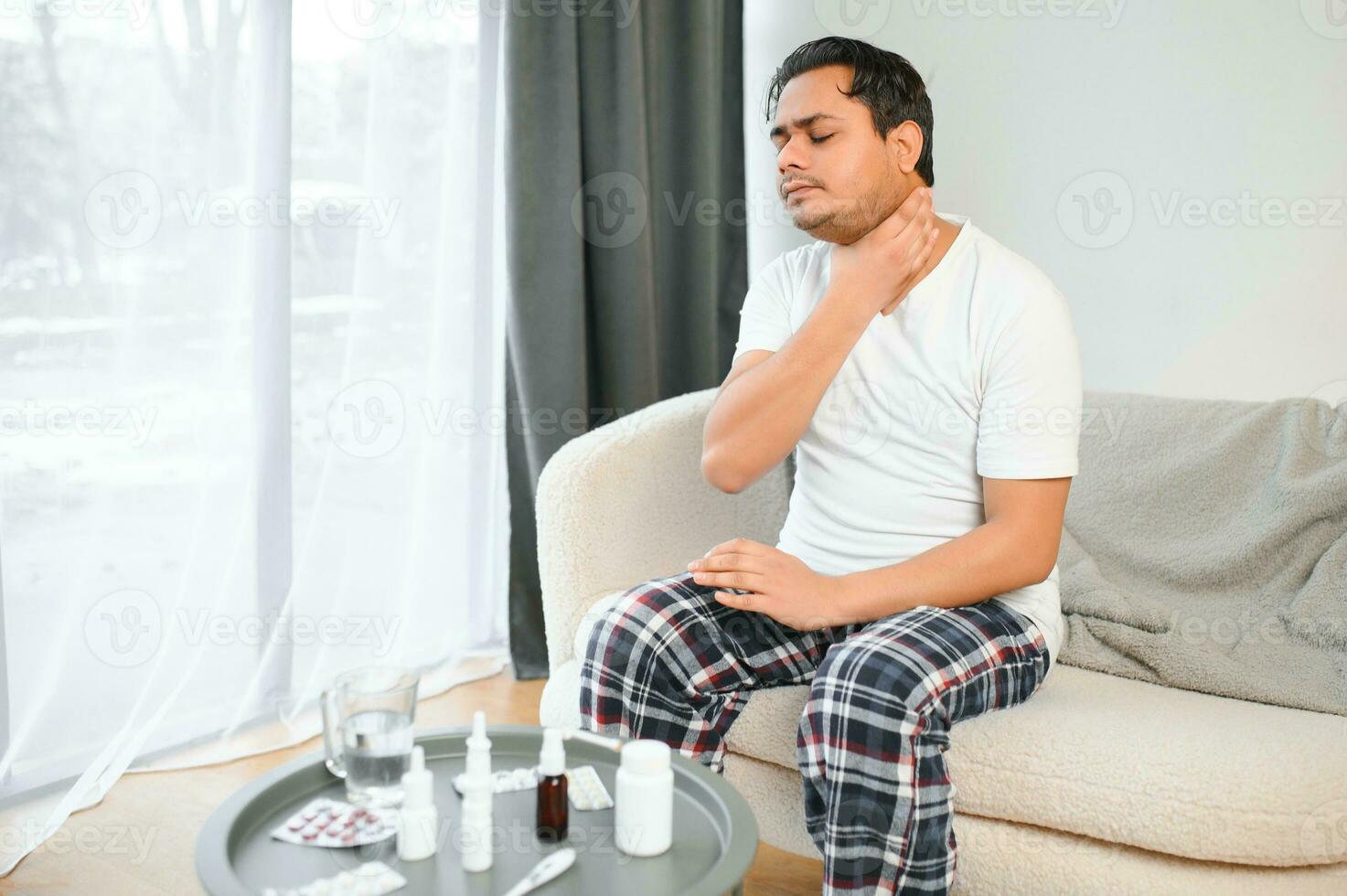 Young Muslim man suffering from a sore throat, sitting on the sofa at home and grimacing, holding his throat. Needs medical treatment photo