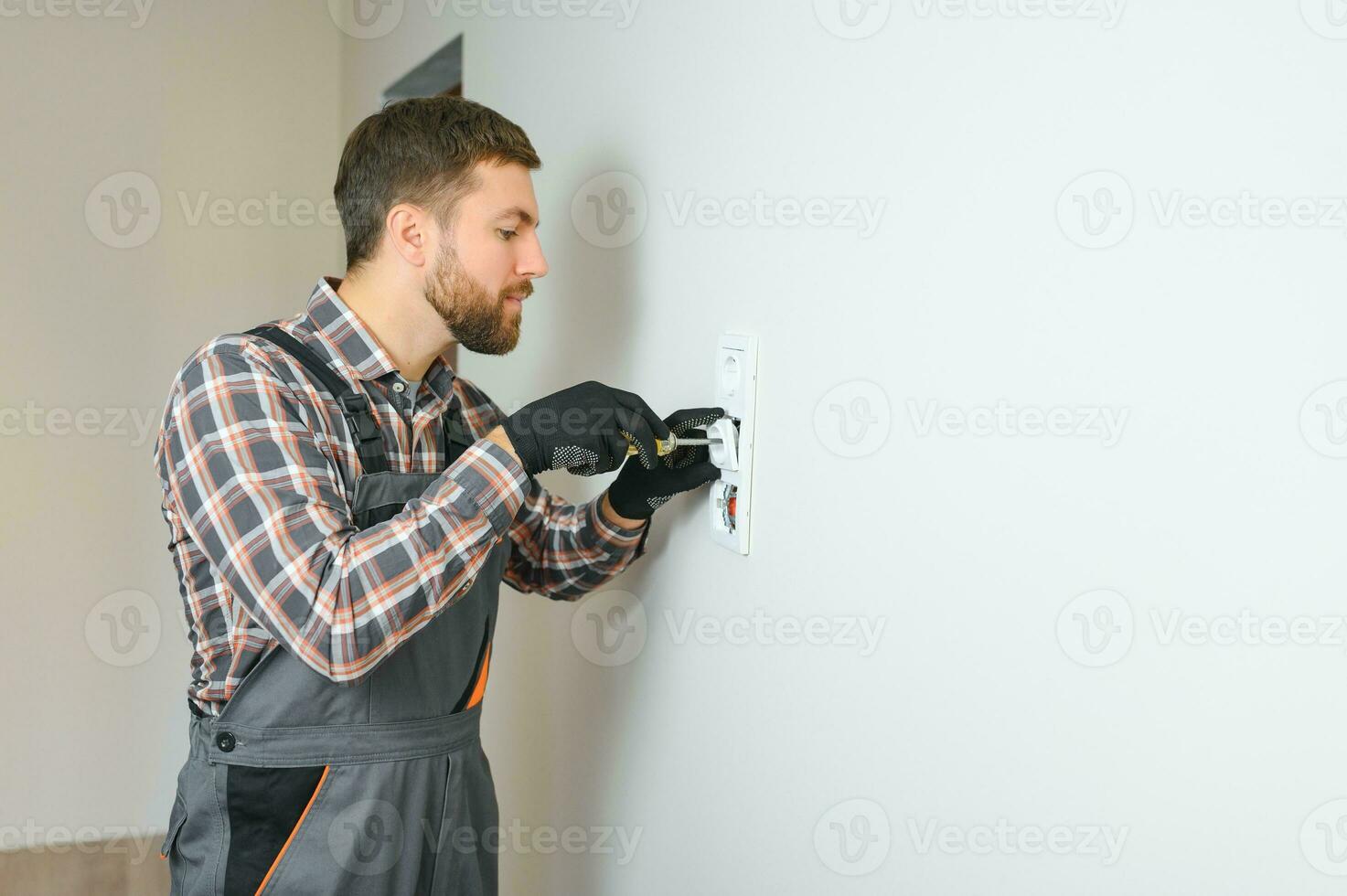 Confident professional electrician in uniform using screwdriver while replacing a socket in apartment after renovation work. photo