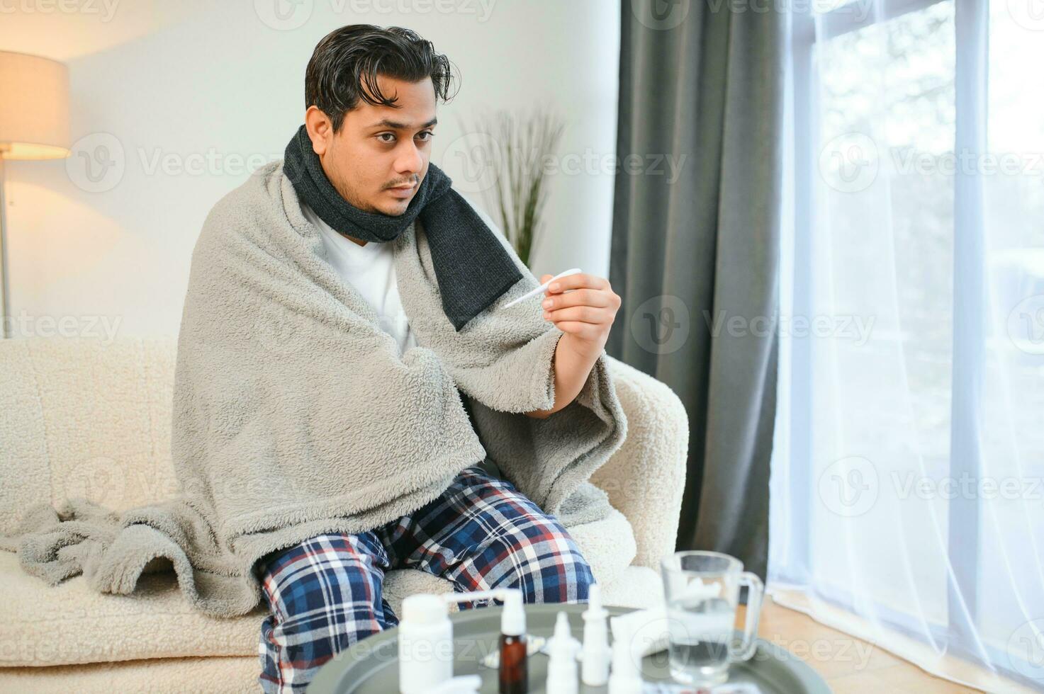 people, health and fever concept - sick indian man in blanket measuring temperature by thermometer at home photo