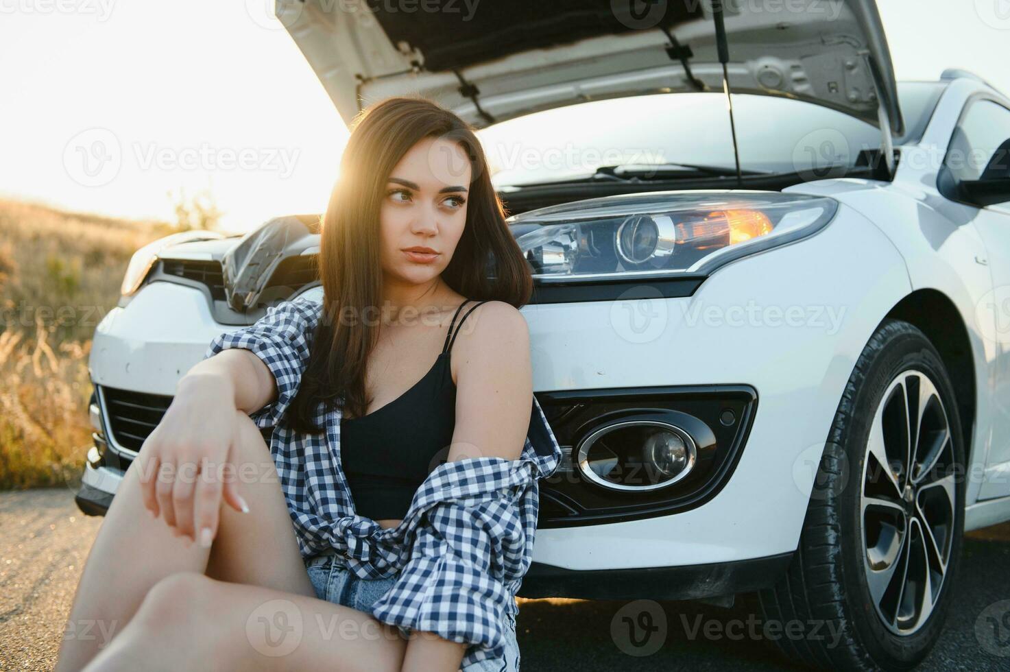 A young girl sits near a broken car on the road with an open hood. photo