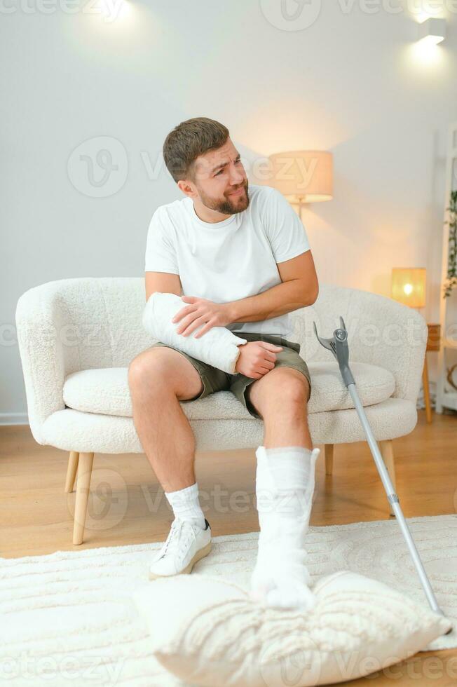 man recovery from accident fracture broken bone injury with leg splints in cast neck splints collar arm splints sling support arm in living room. Social security and health insurance concept photo