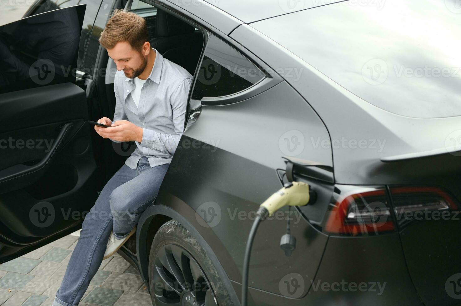 Happy man using smart phone and charging car at vehicle charging station photo