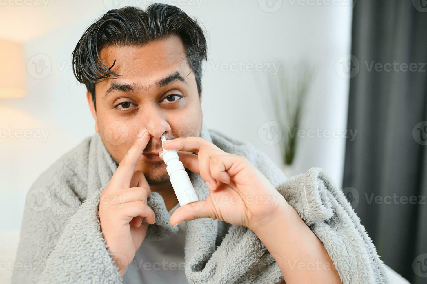 rhinitis, medicine and healthcare concept - sick indian man in blanket using nasal spray at home photo