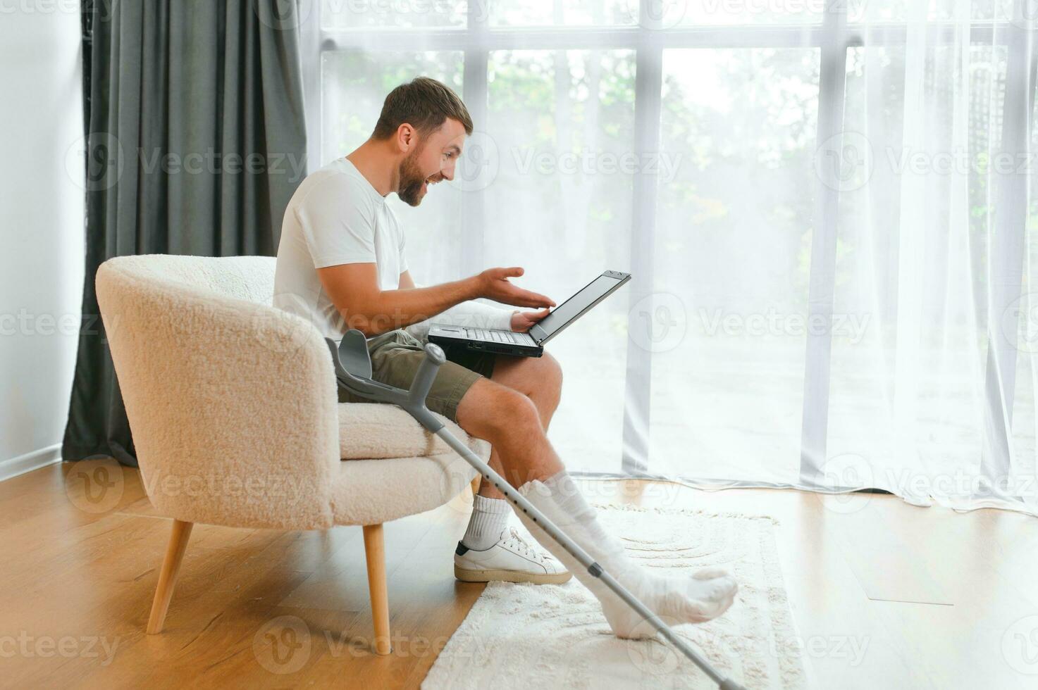 Happy young man with arm in a cast sitting on the couch at home and communicating on a laptop photo