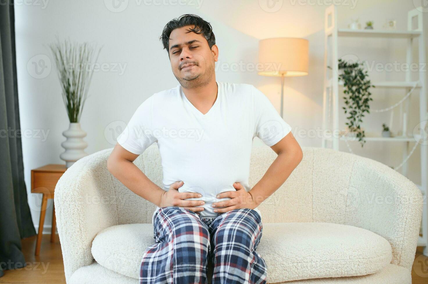 Unhappy indian or arabian man, sits on comfortable sofa in cozy living room, holds his hands on his stomach, grimaces from pain in his stomach, suffers from poisoning, spasm, stomach problems. photo