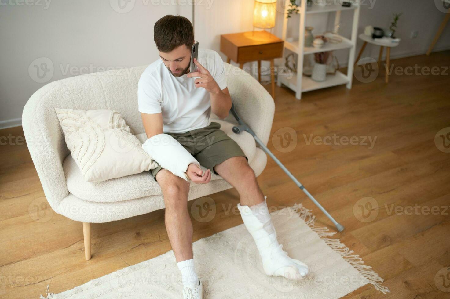 Man with a broken leg and arm using his mobile phone while relaxing on the sofa at home. Accident, injury, treatment, rehabilitation concept photo