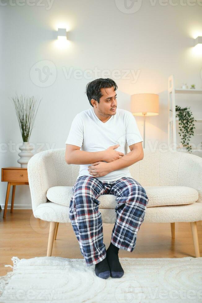 Sad exhausted guy of indian and arabian ethnicity, holds his hands on his stomach, grimaces from pain in his stomach, suffers from poisoning, spasm, stomach problems photo