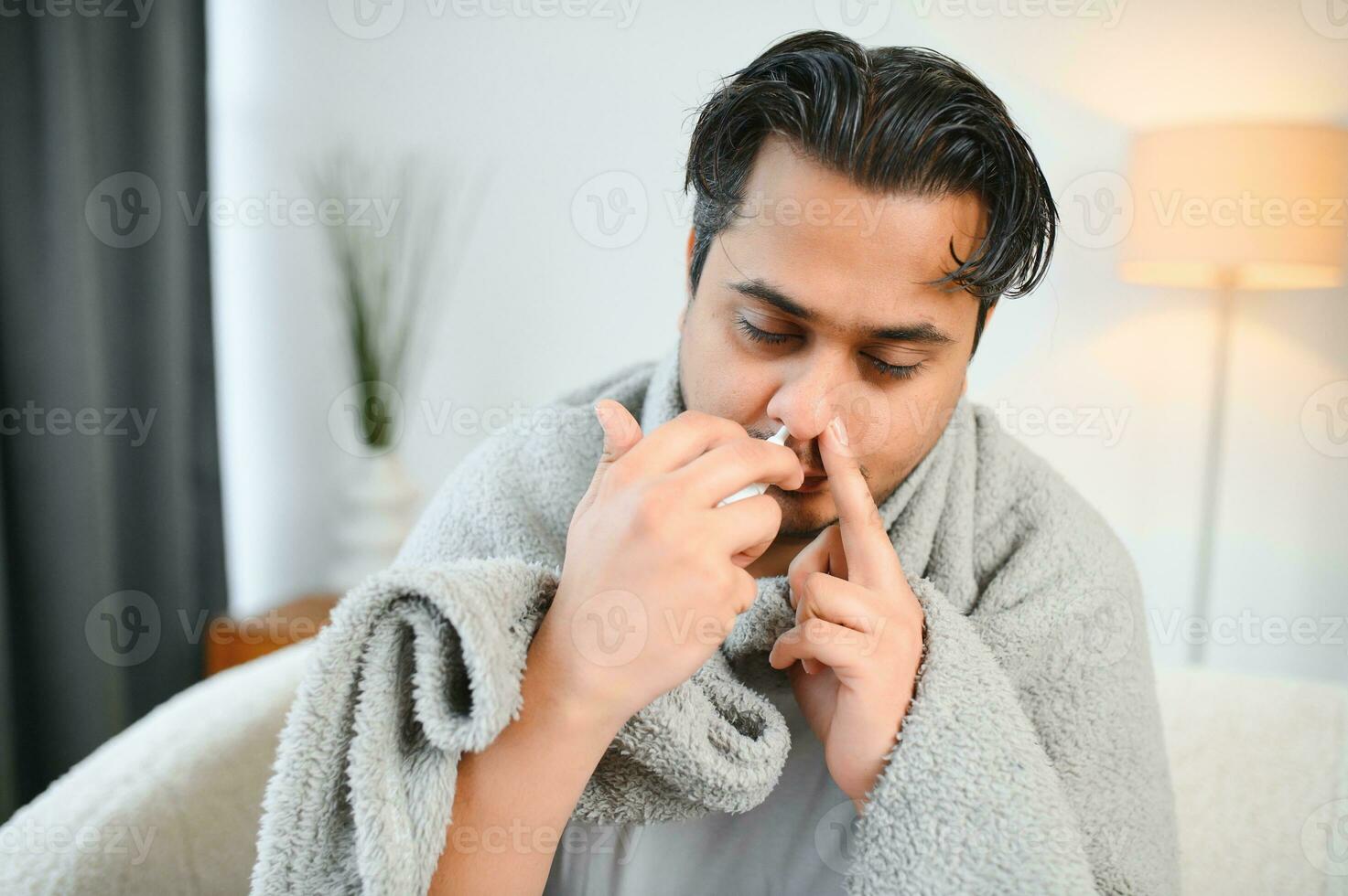 rhinitis, medicine and healthcare concept - sick indian man in blanket using nasal spray at home photo