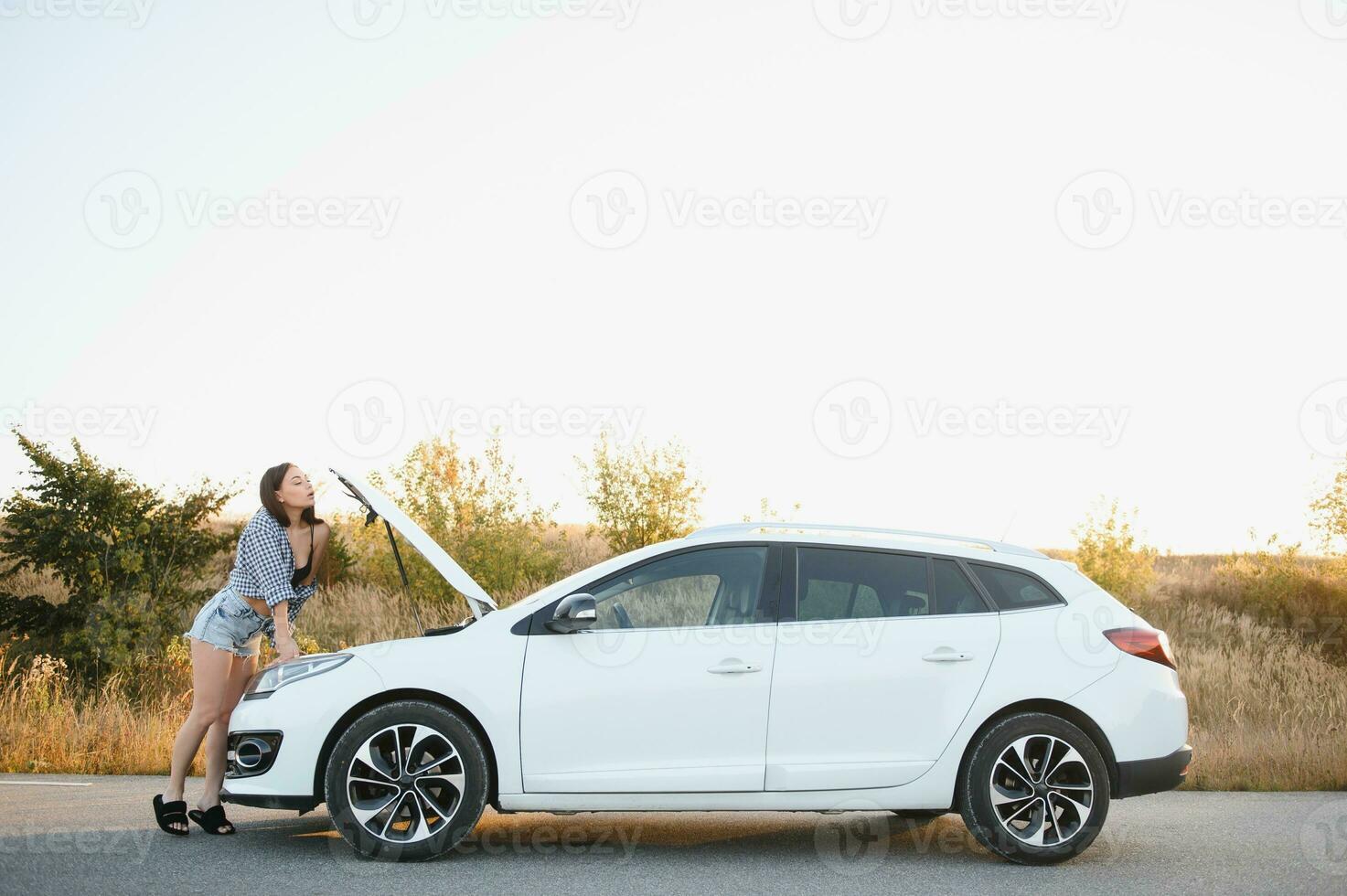 Beautiful slim girl in shirt and shorts looks in open car hood on a road photo