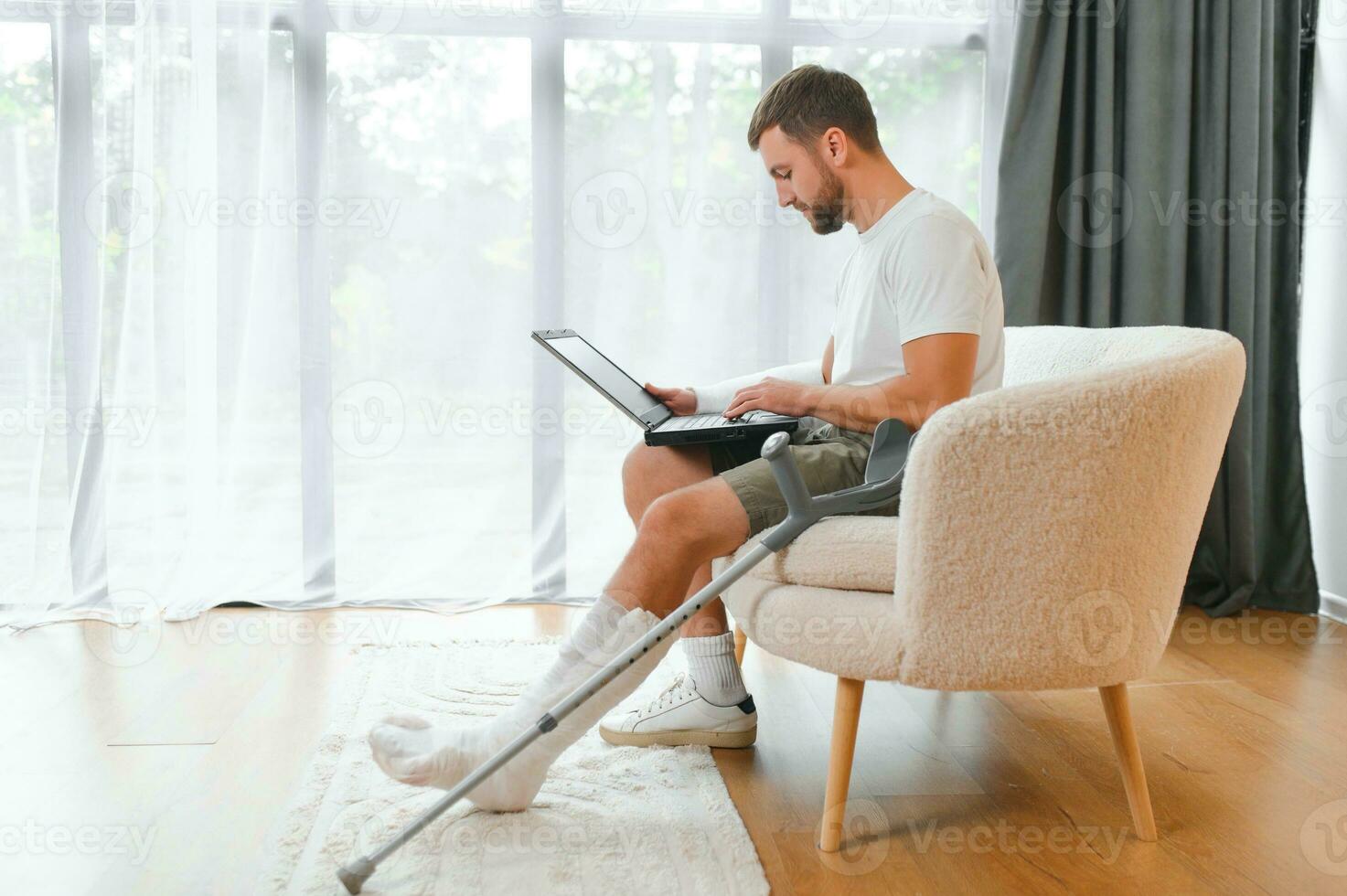 Unhealthy businessman with injured leg and arm in bandage sit on sofa at home work online on computer photo