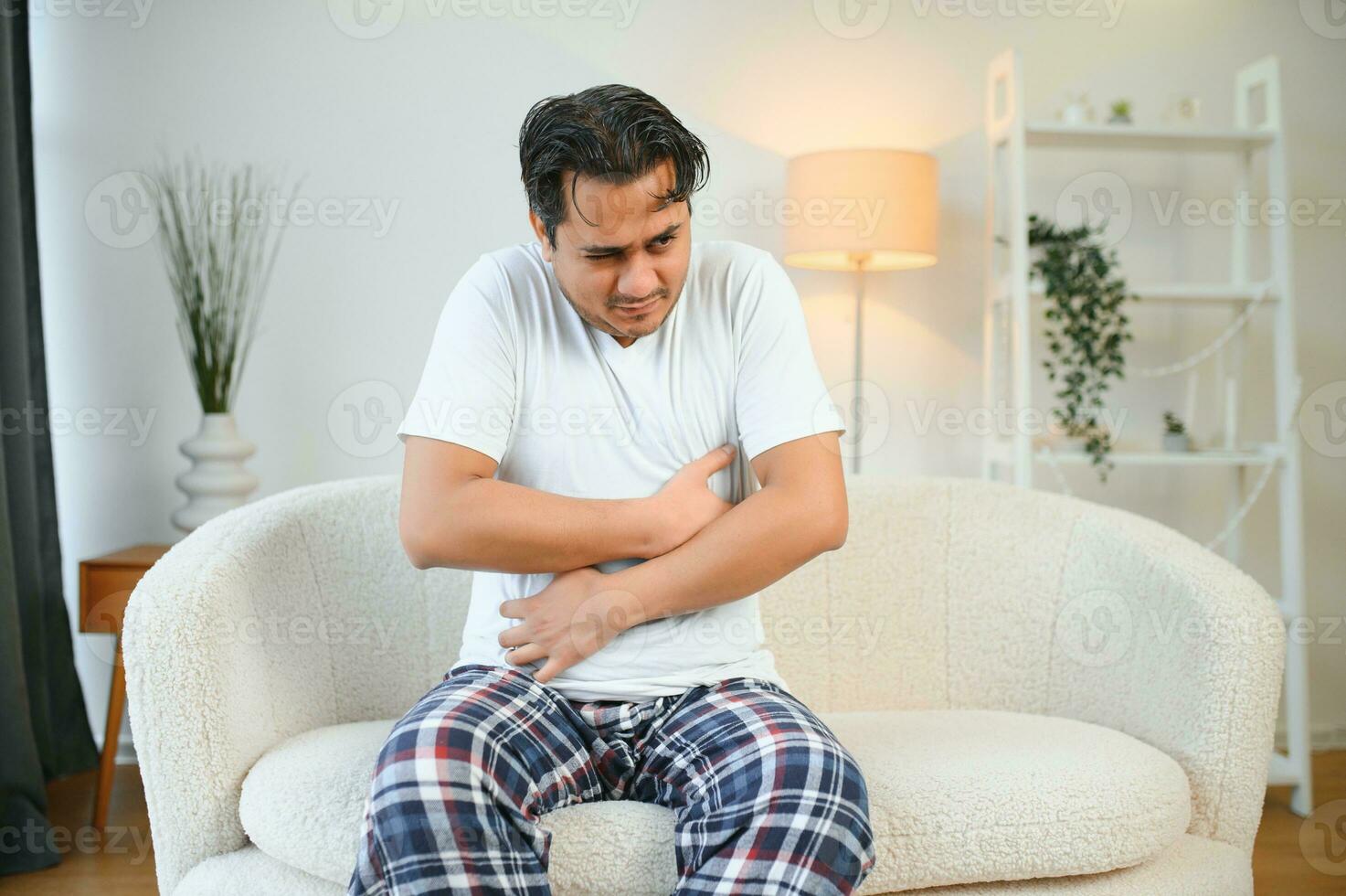 Unhappy indian or arabian man, sits on comfortable sofa in cozy living room, holds his hands on his stomach, grimaces from pain in his stomach, suffers from poisoning, spasm, stomach problems. photo