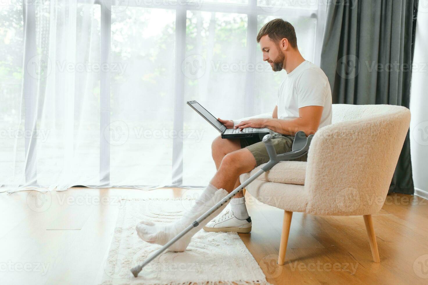 Happy young man with arm in a cast sitting on the couch at home and communicating on a laptop photo