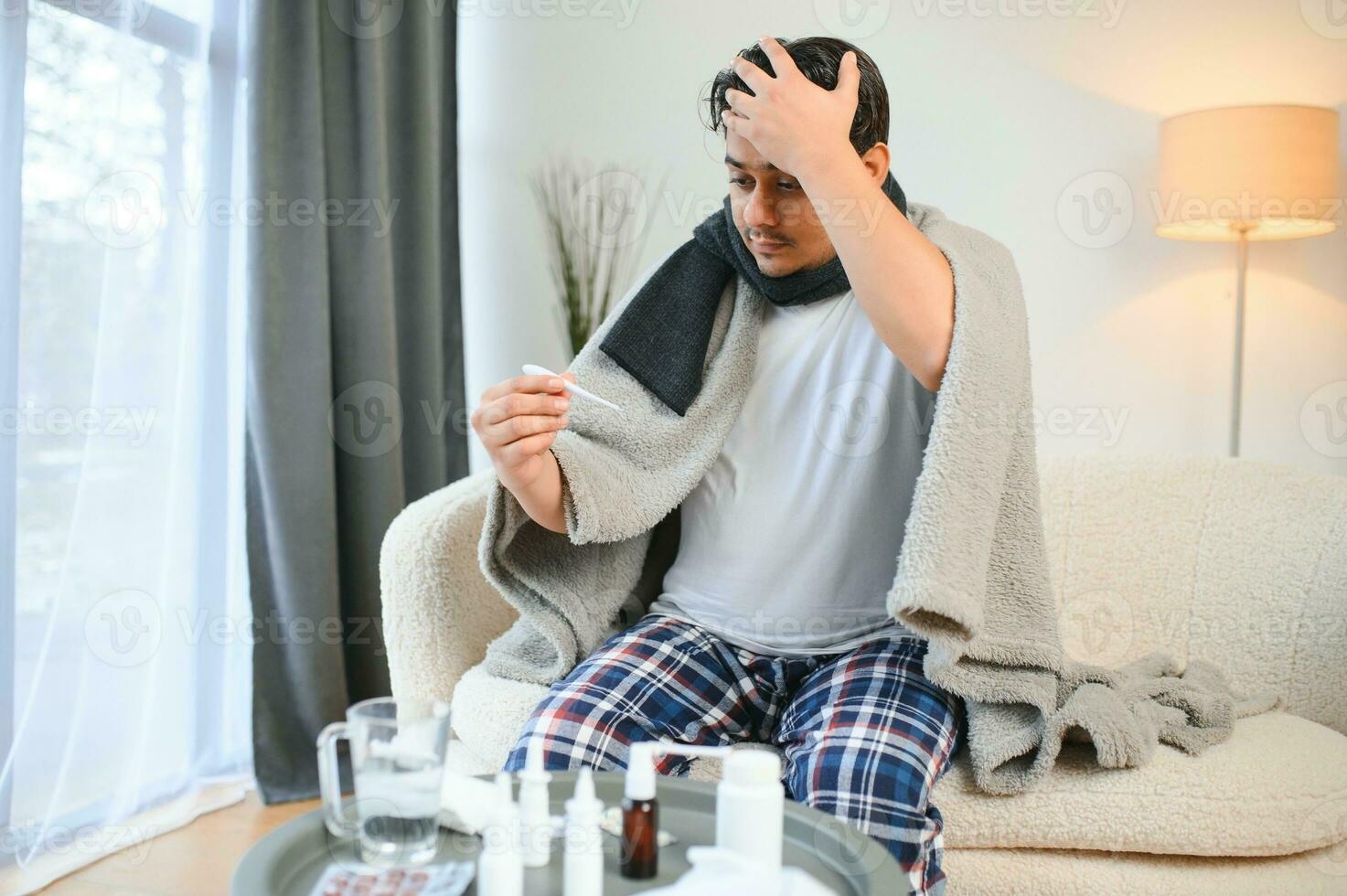 people, health and fever concept - sick indian man in blanket measuring temperature by thermometer at home photo