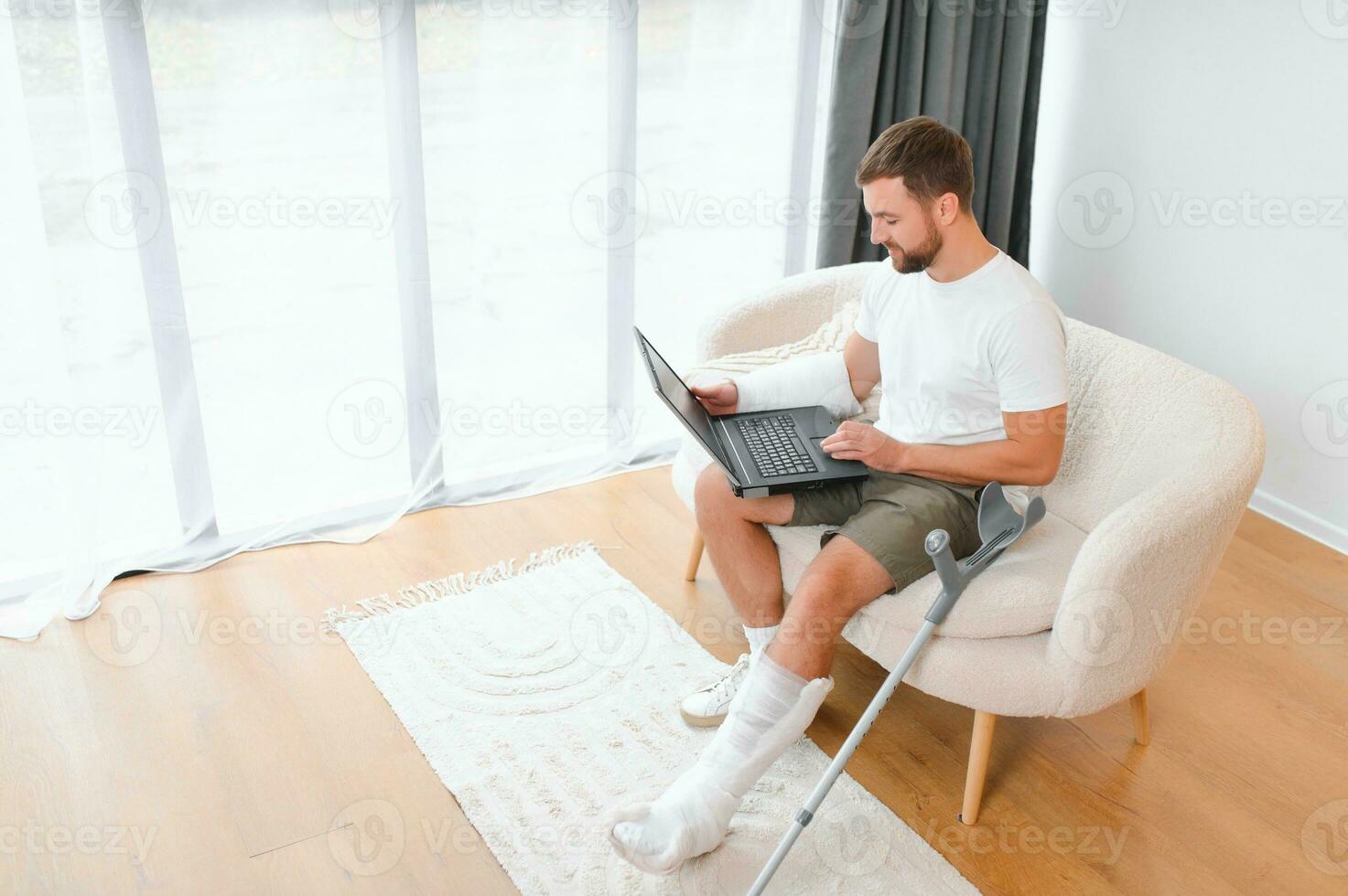 Happy young man with arm in a cast sitting on the couch at home and communicating on a laptop photo