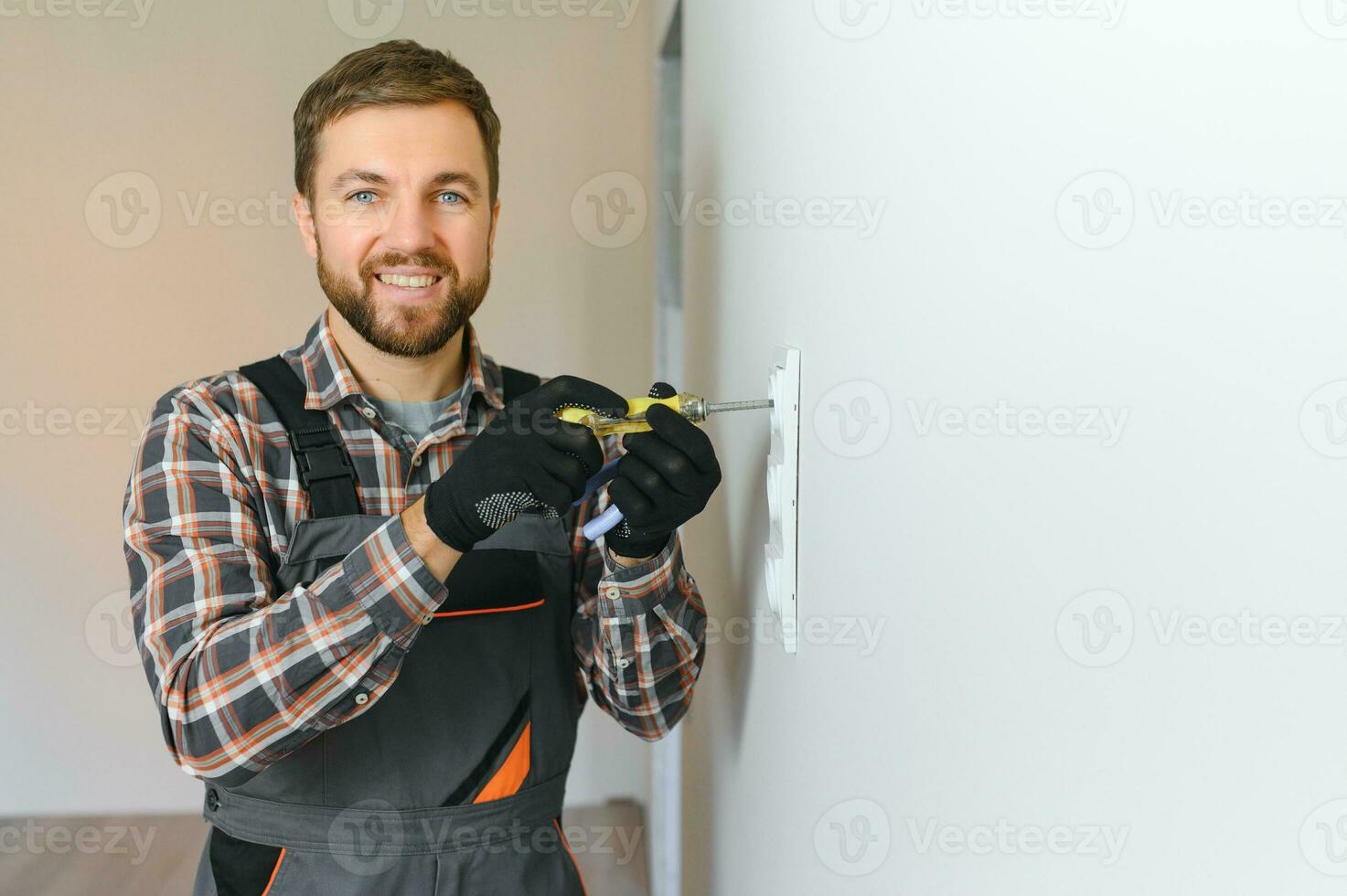 Confident professional electrician in uniform using screwdriver while replacing a socket in apartment after renovation work. photo