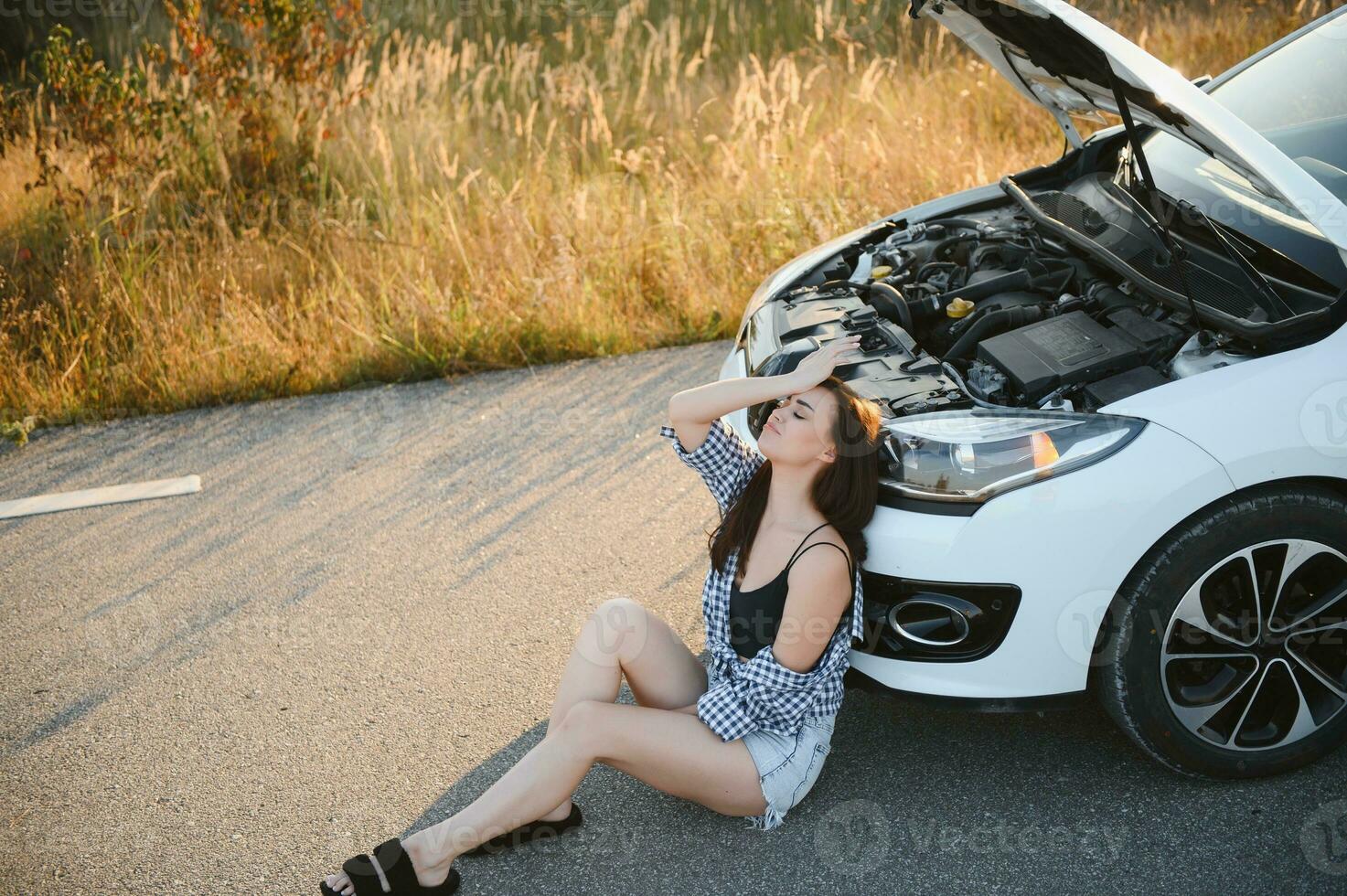 A young girl sits near a broken car on the road with an open hood. photo