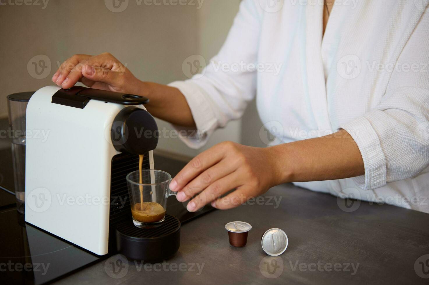 Close-up of a young woman in white bathrobe, making her morning espresso using capsule coffee machine for home use photo